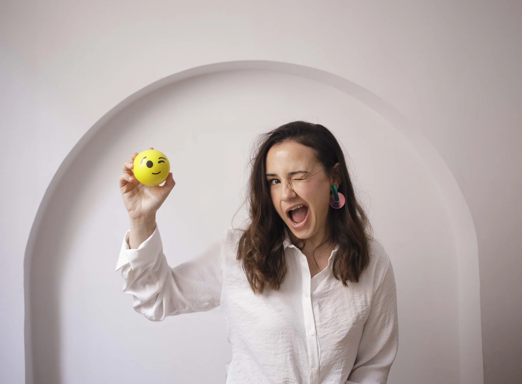 Lidia Viso con cabello castaño y blusa blanca, haciendo una expresión divertida con un ojo cerrado, sosteniendo una pelota amarilla con un emoji de guiño y sonrisa.