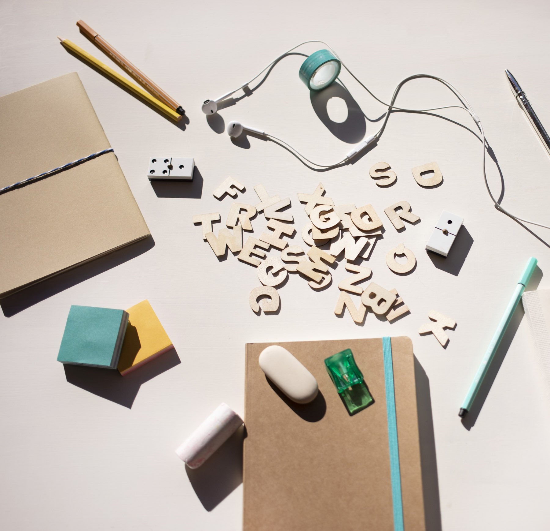 objetos de papelería sobre una mesa, incluyendo un cuaderno de color beige, notas adhesivas de varios colores, una caja de fósforos, letras de madera formando las palabras 'TRABAJO' y otras letras dispersas, auriculares blancos, cinta adhesiva de color verde, una libreta pequeña, pegamento en barra y un bolígrafo