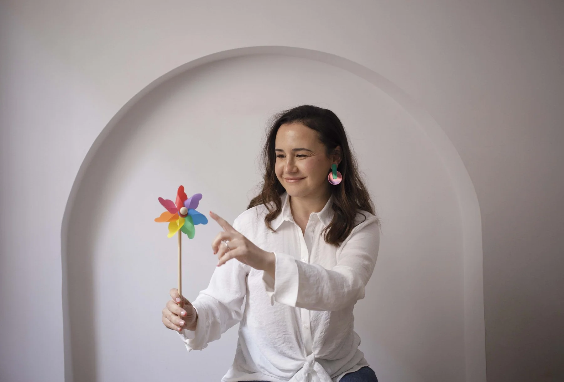Lidia Viso sonriente usando camiseta blanca, sosteniendo una veleta de colores brillante y un pendiente grande, en un fondo con forma de arco en pared blanca.