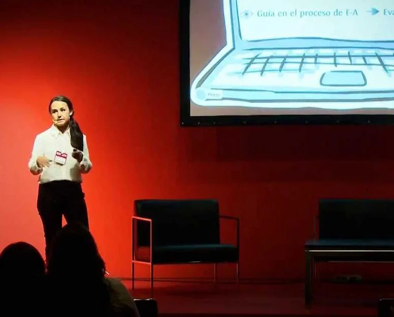 Lidia Viso de pie en un escenario, hablando, con un fondo rojo y una pantalla de proyector que muestra un dibujo de una computadora portátil.