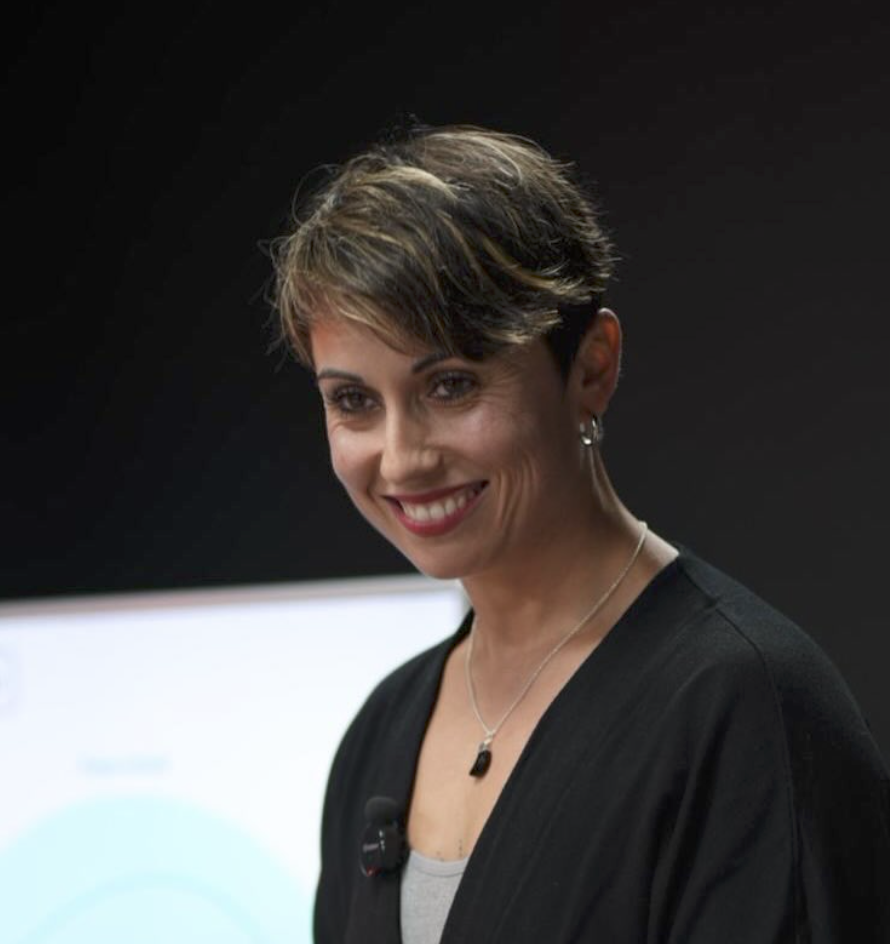 Mujer con cabello corto, vestida con ropa negra, sonriendo en un ambiente oscuro.
