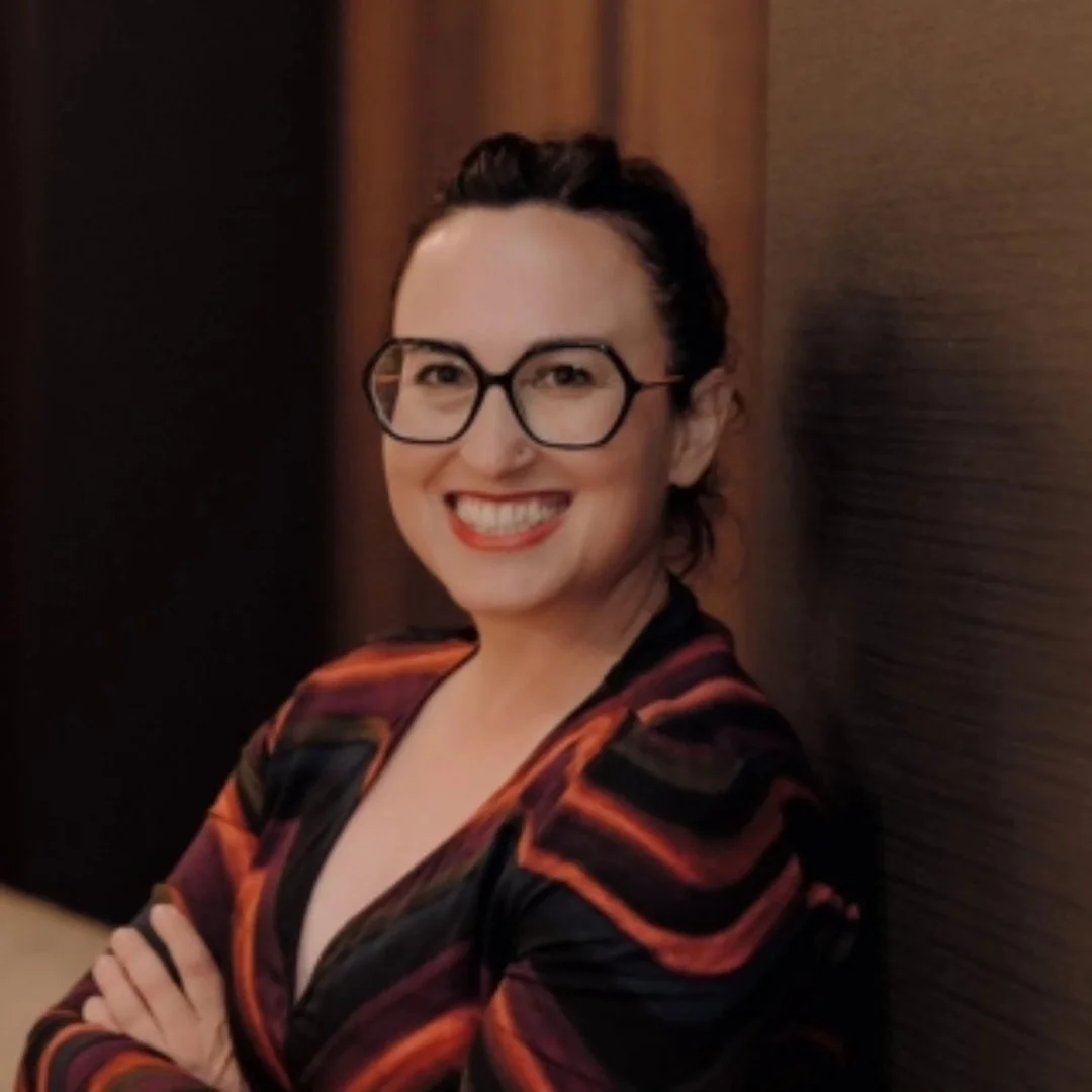 Mujer sonriendo, con gafas y vestido con patrones en tonos oscuros y naranjas, de cabello oscuro peinado hacia atrás, en un entorno con paredes de tonos marrón.