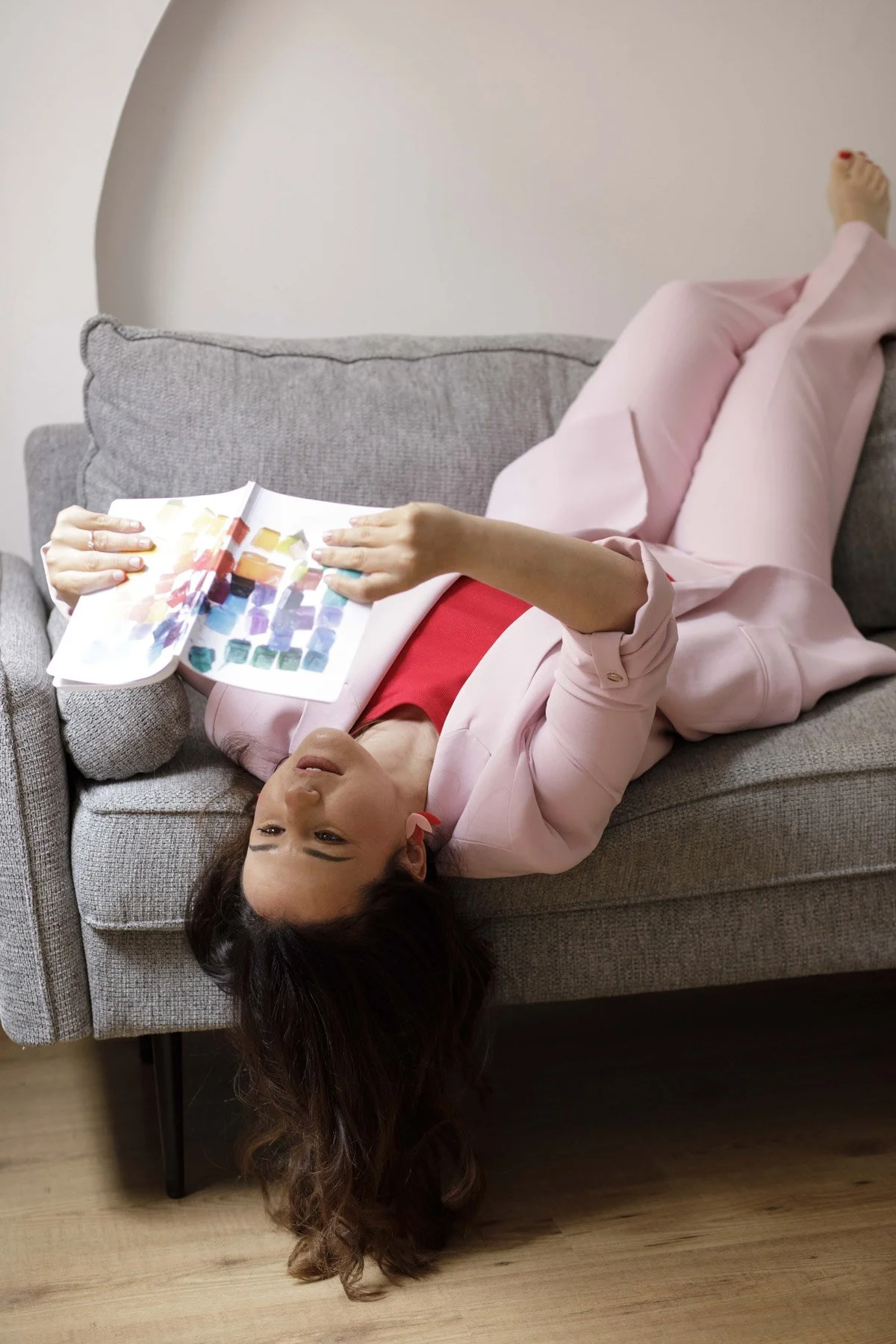 Lidia Viso acostada en un sofá, leyendo un libro con colores y patrones, con cabello oscuro y piel clara, vestida con blusa rosa y pantalones rosas, con un piso de madera.