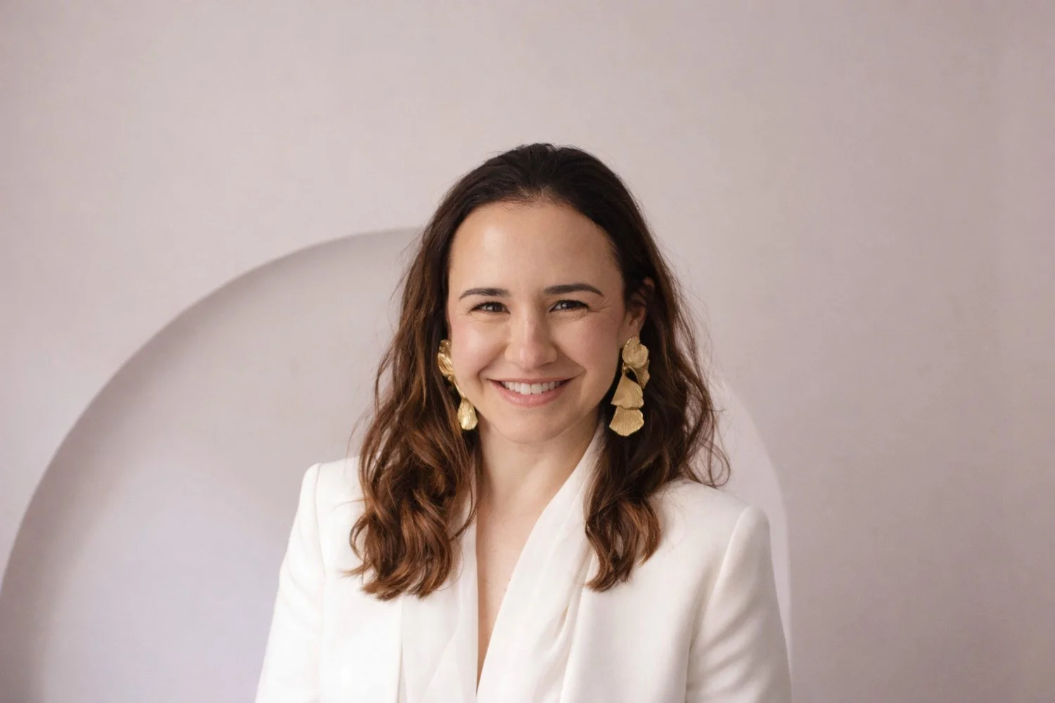 Retrato de Lidia Viso sonriendo, con bigote y aretes grandes dorados, viste una blusa blanca y está frente a un fondo neutro con formas geométricas