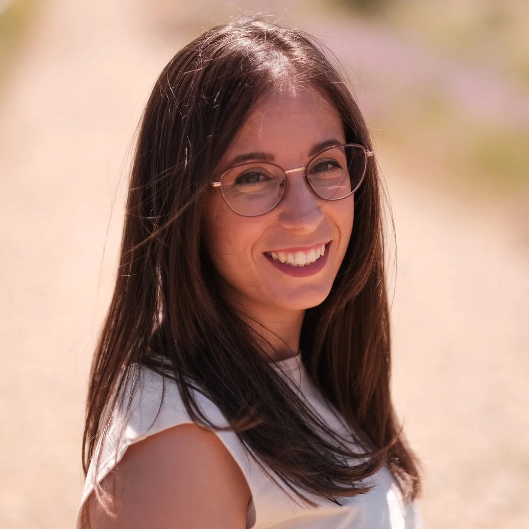 Mujer joven con gafas, cabello largo y castaño, sonriendo en un fondo difuso y soleado.