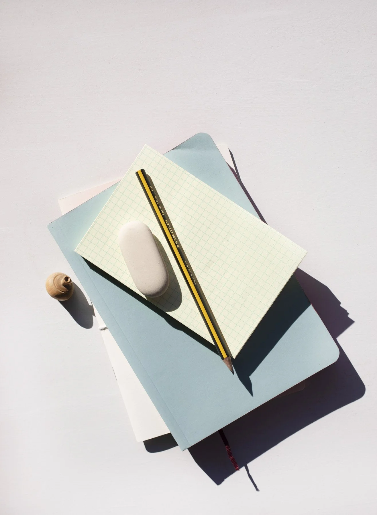 Varios cuadernos apilados en tonos claros, acompañados de un bolígrafo negro y amarillo, una goma de borrar y una pequeña concha en una superficie blanca con sombra suave.