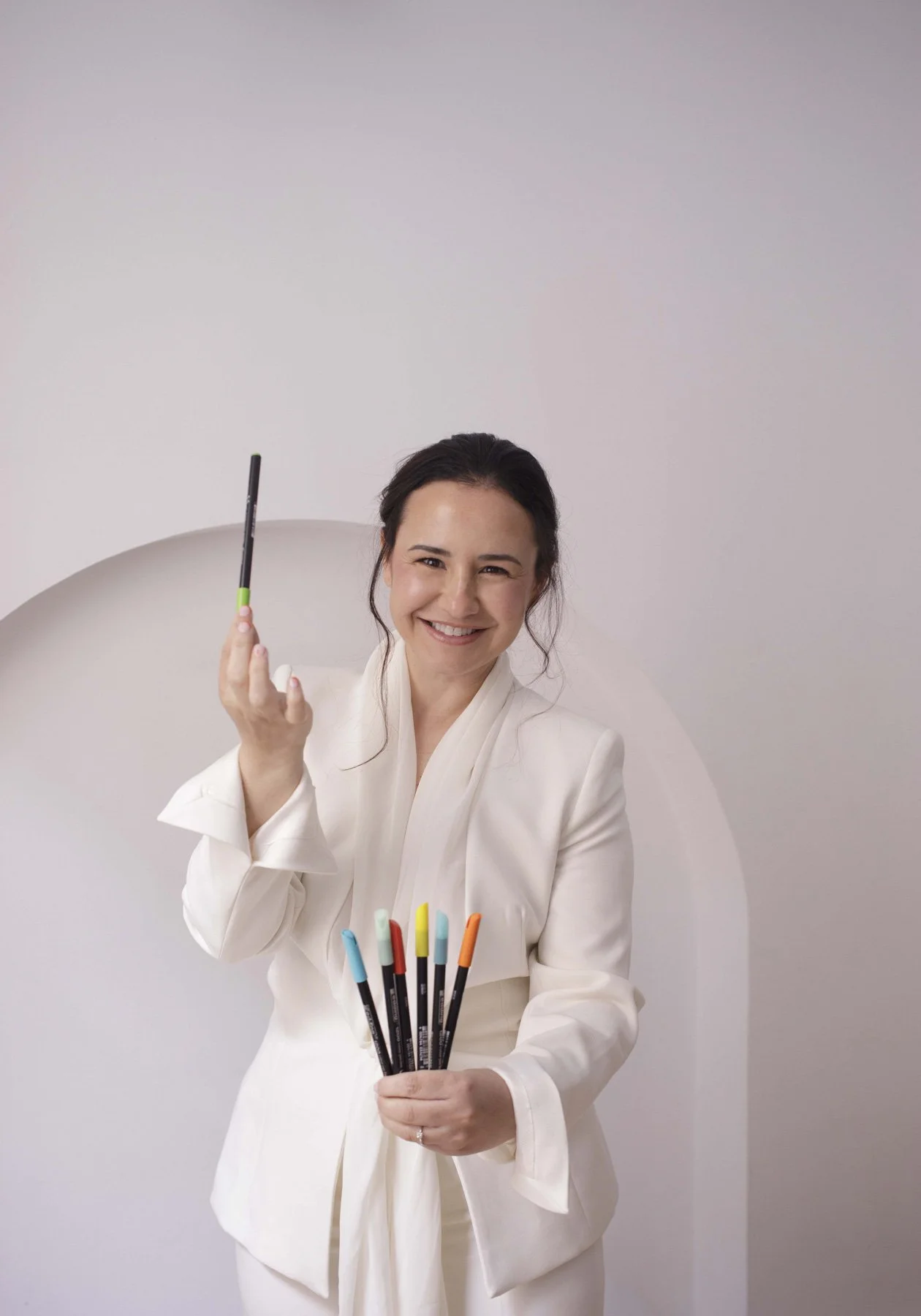 Lidia Viso sonriendo, sosteniendo un bolígrafo en una mano y varias plumas de colores en la otra, en un fondo claro.
