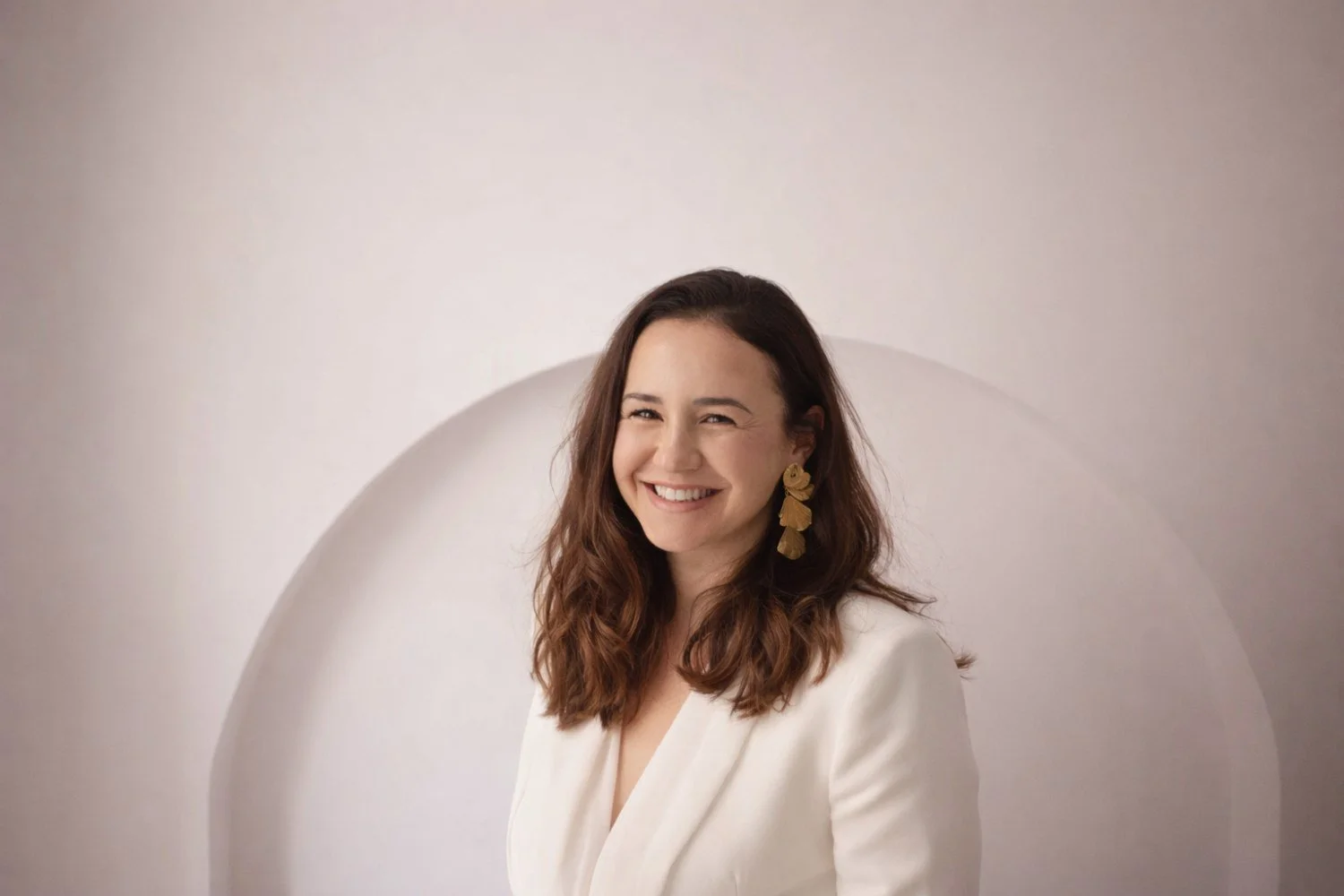 Lidia Viso con cabello castaño oscuro, vestido blanco y pendientes dorados, sonriendo y mirando a la cámara, fondo neutro con formas circulares.
