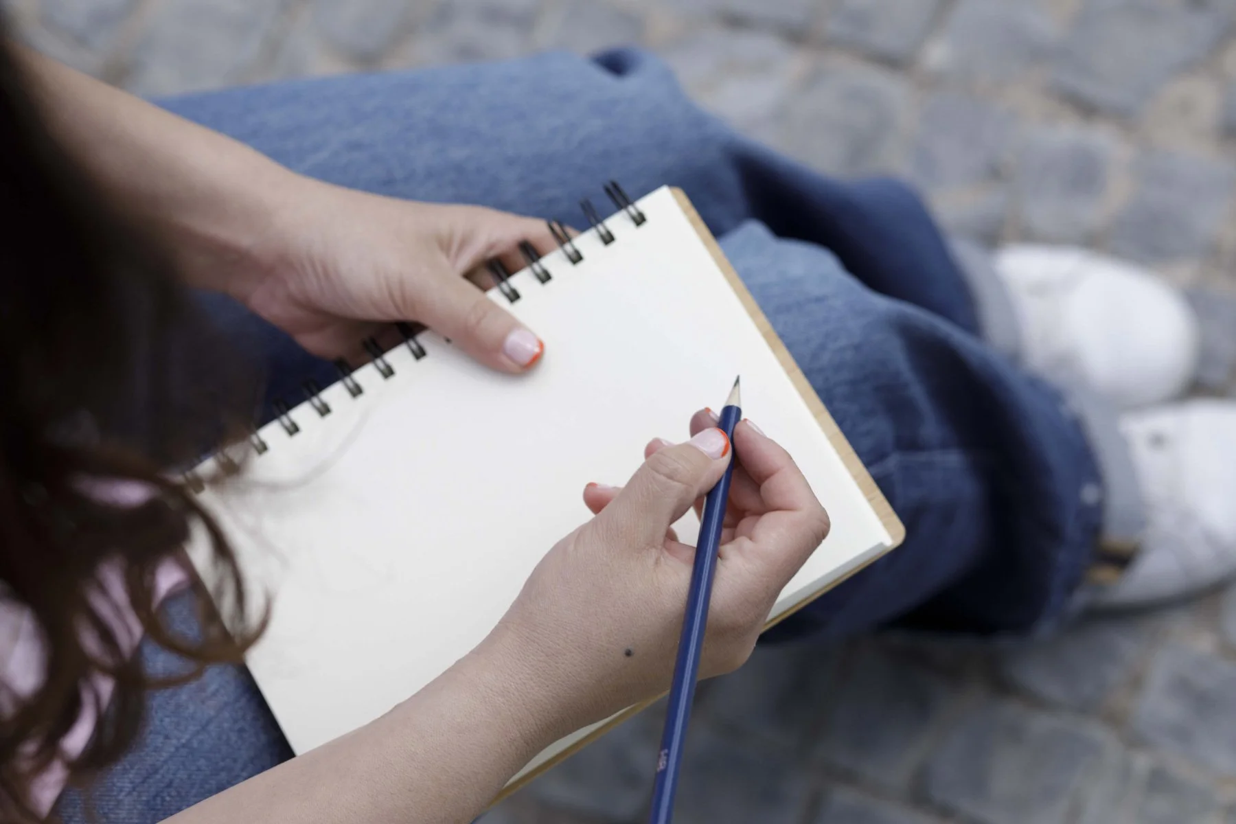 Persona sentada en una banca escribiendo en un cuaderno con lápiz, vista parcial de sus piernas y fondo de piedra.