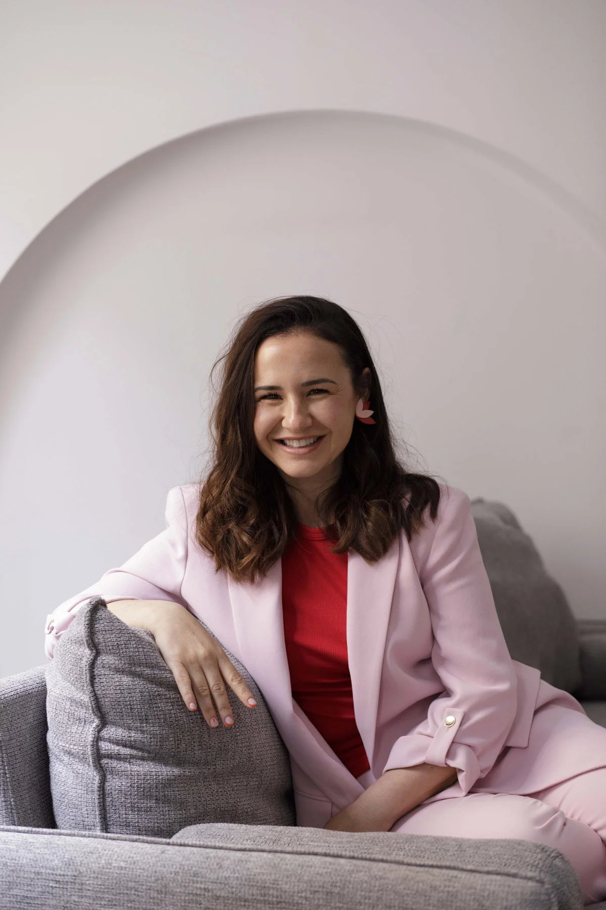 Lidia Viso sonriendo, sentada en un sillón gris, usando ropa rosa y roja, con pendientes de corazones rosados y rojos
