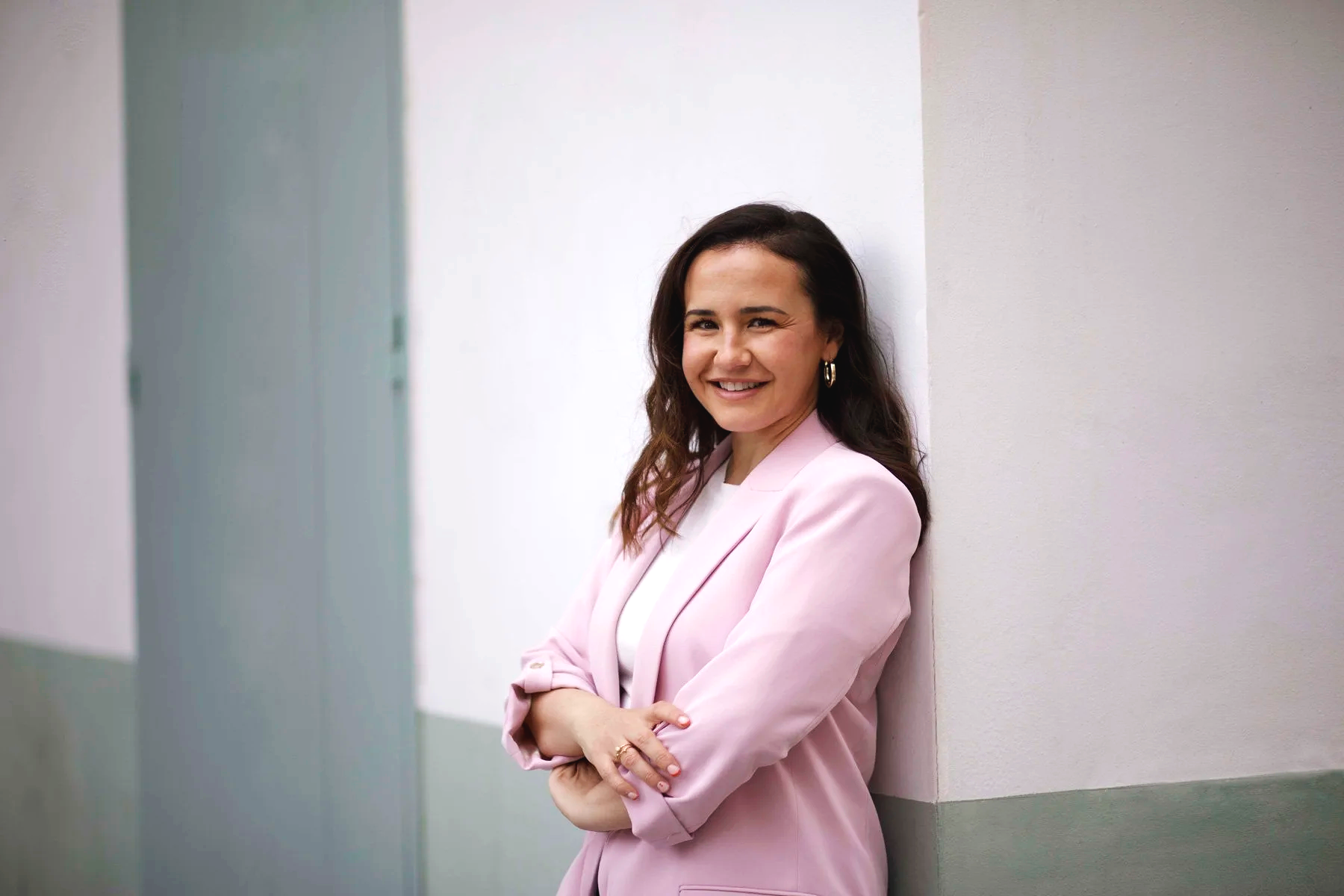 Lidia Viso sonriendo, con brazos cruzados, vestida con saco rosa claro, apoyada en la pared blanca, en un pasillo con paredes blancas y verdes.