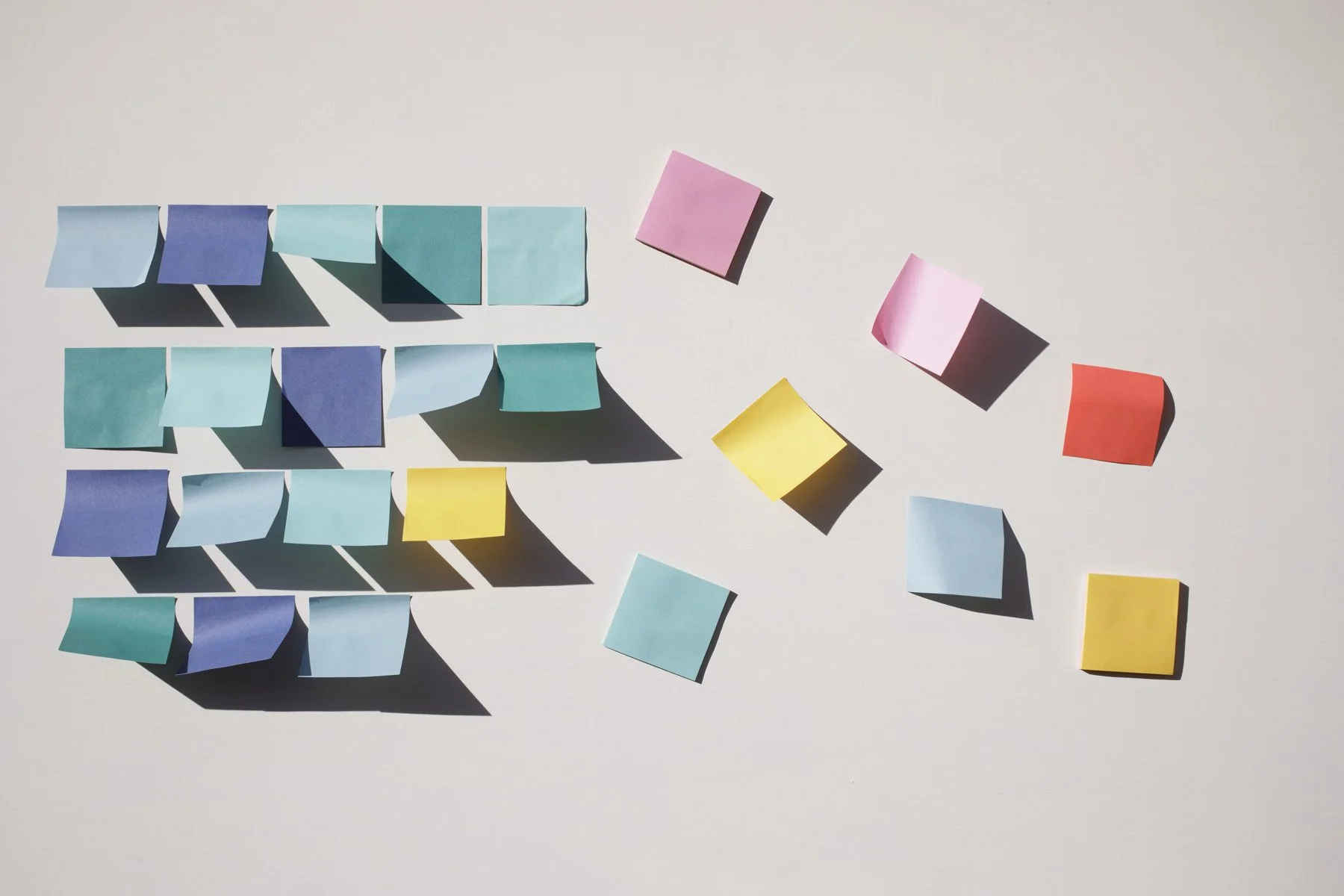 Hoja de papel de notas de colores en una superficie, algunas pegadas y otras cayendo, con sombras proyectadas.