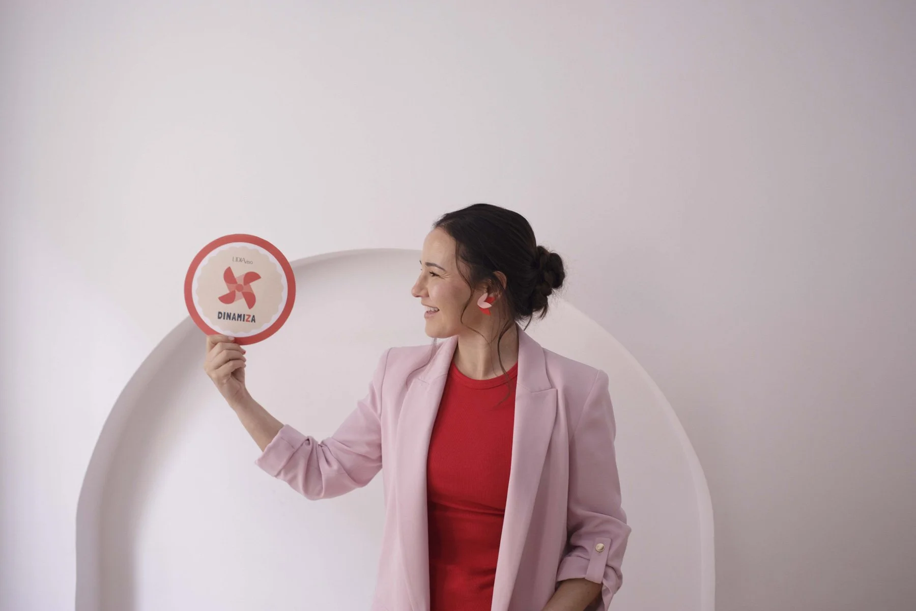Lidia Viso sonriendo, usando blazer rosa y camiseta roja, sosteniendo un cartel redondo con un logo de rotor en color rojo, con la palabra 'DINAMIZA' debajo, en un fondo liso y claro.