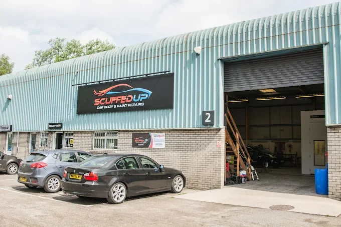 Exterior of a car repair shop named 'Scuffed Up' with a few parked cars outside and a large open garage door.