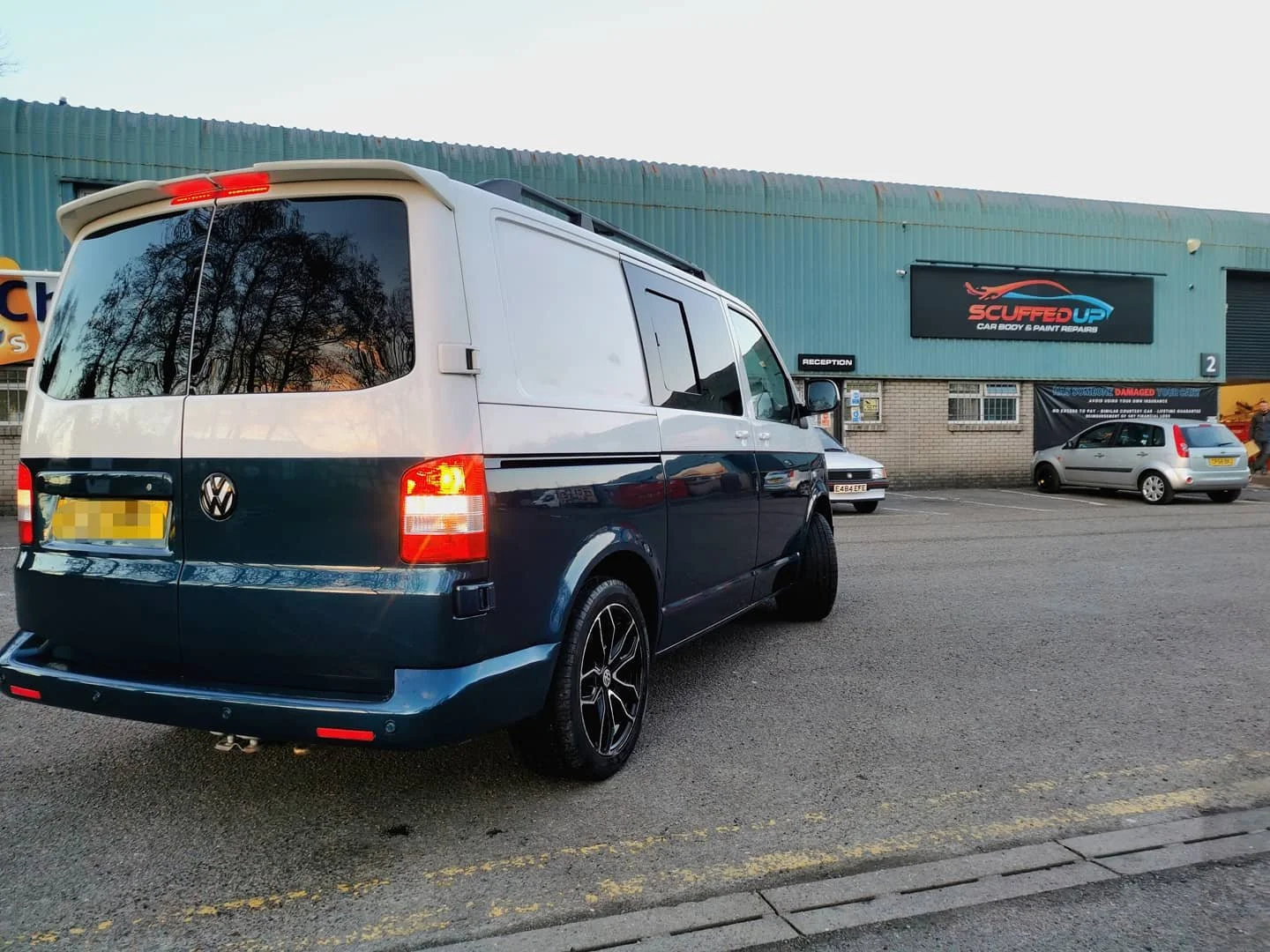 A dark blue Volkswagen van parked in a parking lot outside a car repair shop with a sign reading 'Scuffed Up Car Body & Paint Repairs'. Trees are reflected in the van's rear window and other cars are visible in the background.
