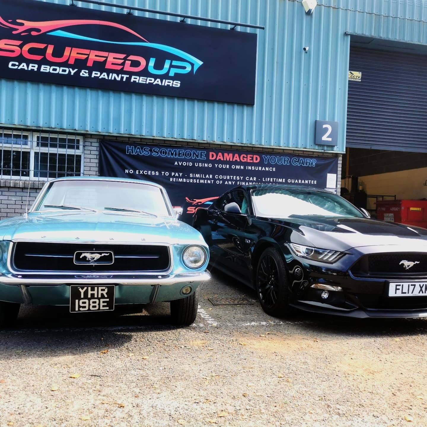 A vintage light blue Ford Mustang car and a modern black Ford Mustang car parked outside a blue industrial building with signs for a car body repair shop - Scuffed Up