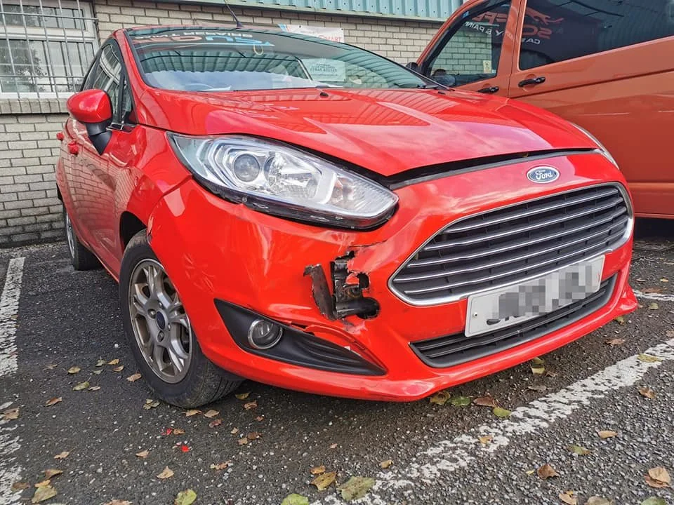 Red Ford Fiesta with front-end damage, missing front bumper and damaged headlight on the driver's side, parked on a street with fallen leaves.