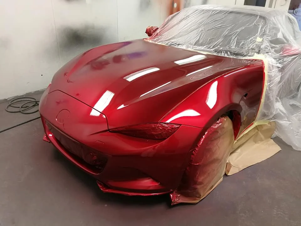 A red sports car being painted or wrapped, partially covered with plastic and masking tape, in a paint booth or workshop.