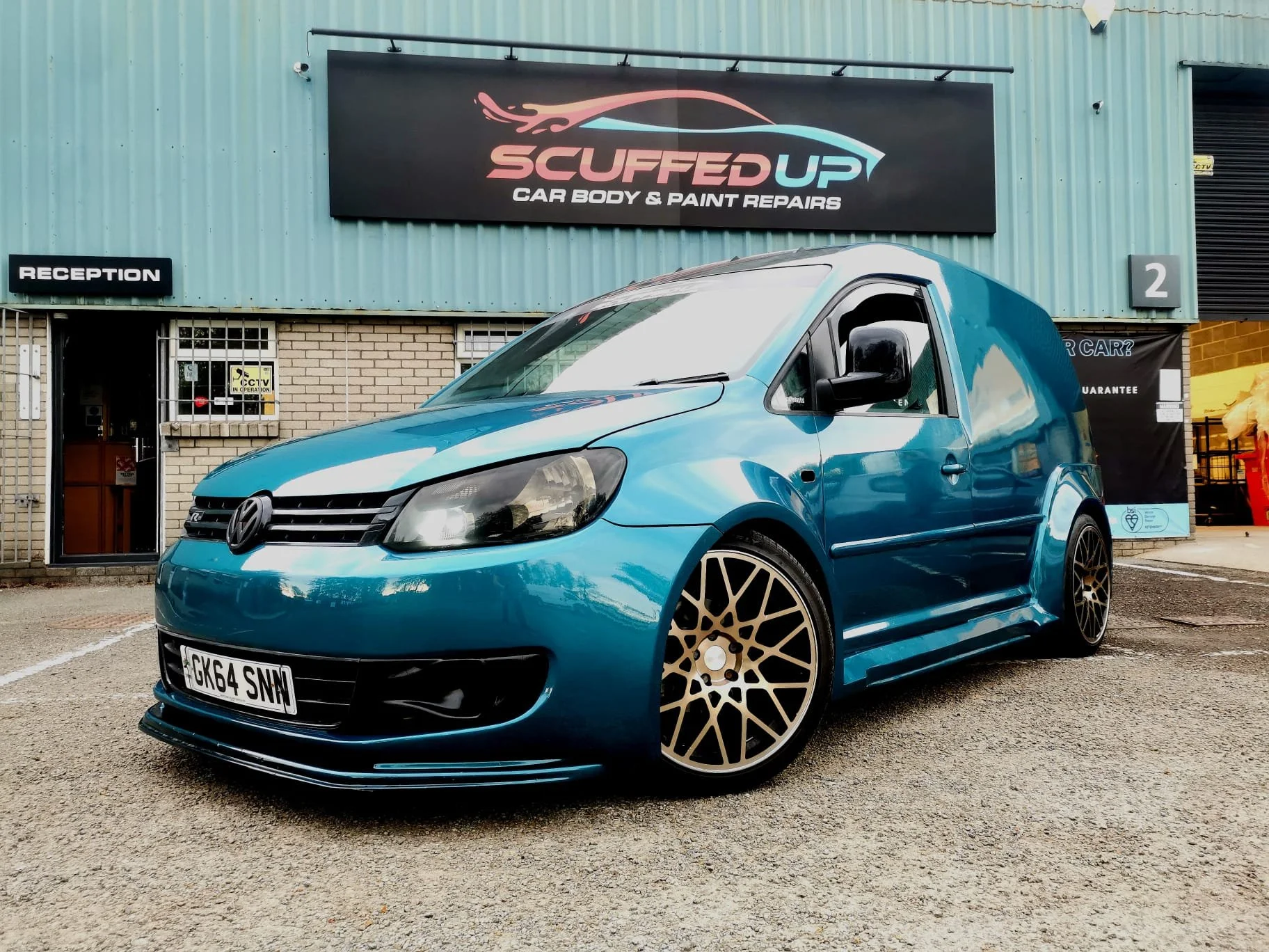 A blue Volkswagen car parked outside a car repair shop, 'Suffer Up Car Body & Paint Repairs', with custom gold alloy wheels.
