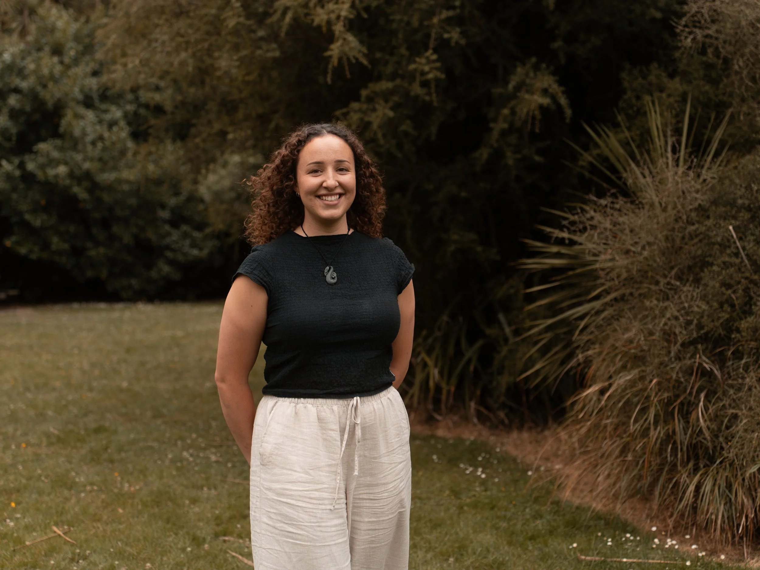 A young woman with curly hair, wearing a black shirt and beige pants, smiling outdoors in a park or garden with trees and bushes in the background.