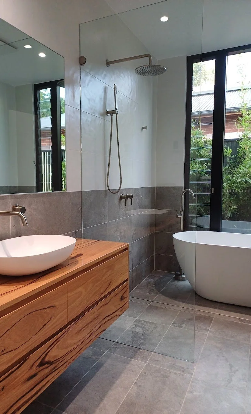 A modern bathroom with a wooden vanity, a white vessel sink, a large window, a freestanding bathtub, and a walk-in shower with a rain showerhead and handheld shower, separated by a glass panel.