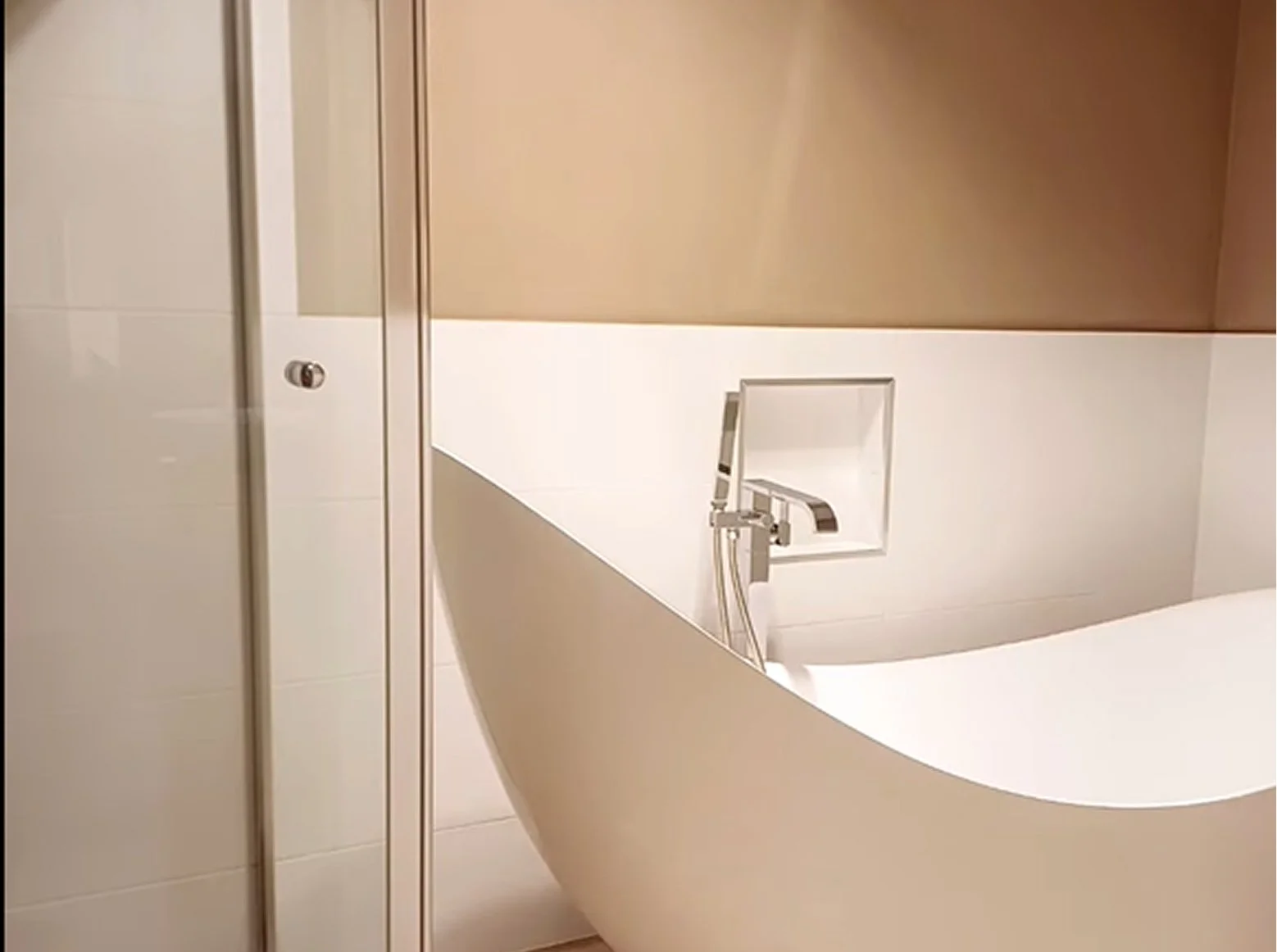 A modern white bathtub with a built-in faucet and a handheld showerhead, set against beige and white tiled walls.
