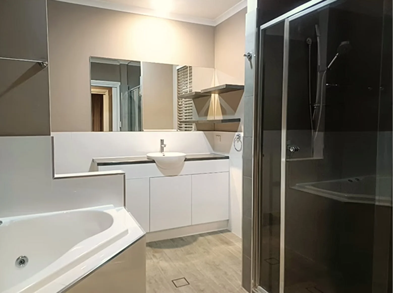 Modern bathroom with large mirror, white vanity with built-in sink, black countertop, and beige walls. There is a corner shower with glass doors on the right and a white whirlpool tub partially visible on the left.