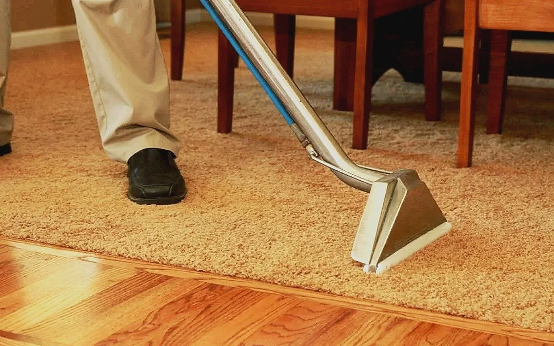 A person in beige pants and black shoes vacuuming a carpeted area next to a wooden floor with a commercial carpet cleaner.