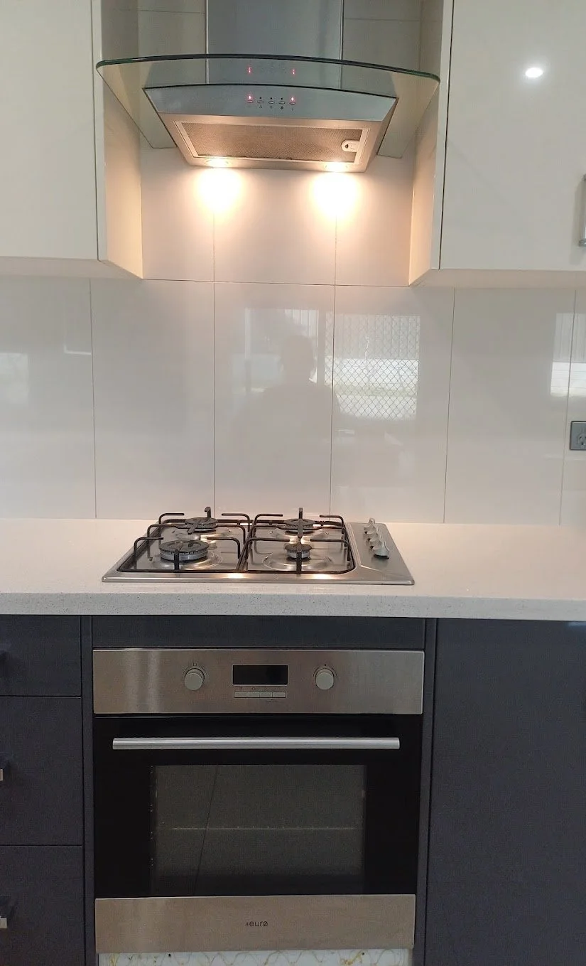 A modern kitchen with a white countertop, gas stove with four burners, and a built-in oven. An under-the-hood range vent is above the stove, with lights on.