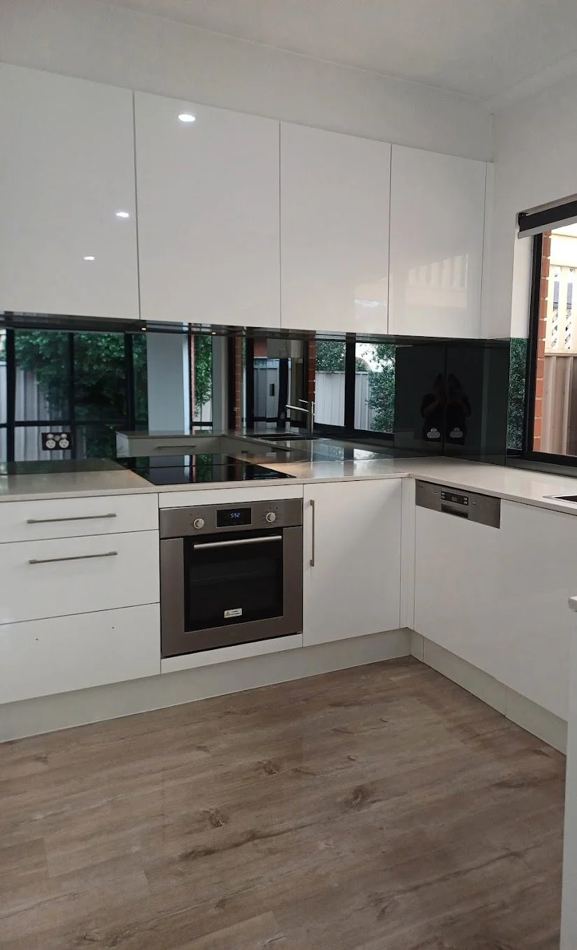 Modern kitchen with white cabinets, a built-in oven, a stovetop, and a dishwasher. Large windows reflect the kitchen interior and outdoor greenery.