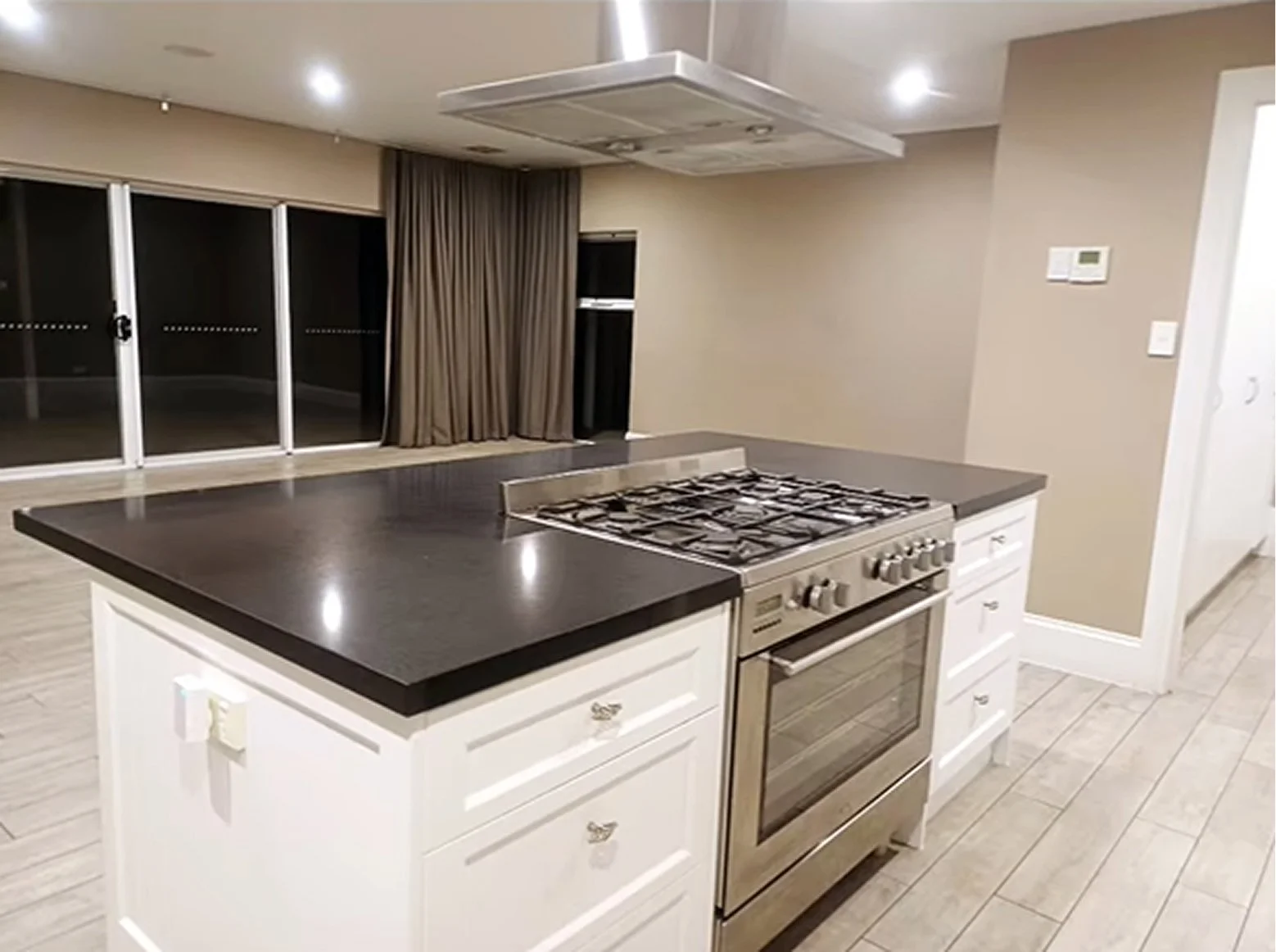 Modern kitchen with a stainless steel oven and black countertop island, connected to a spacious living area with large sliding glass doors and beige curtains.