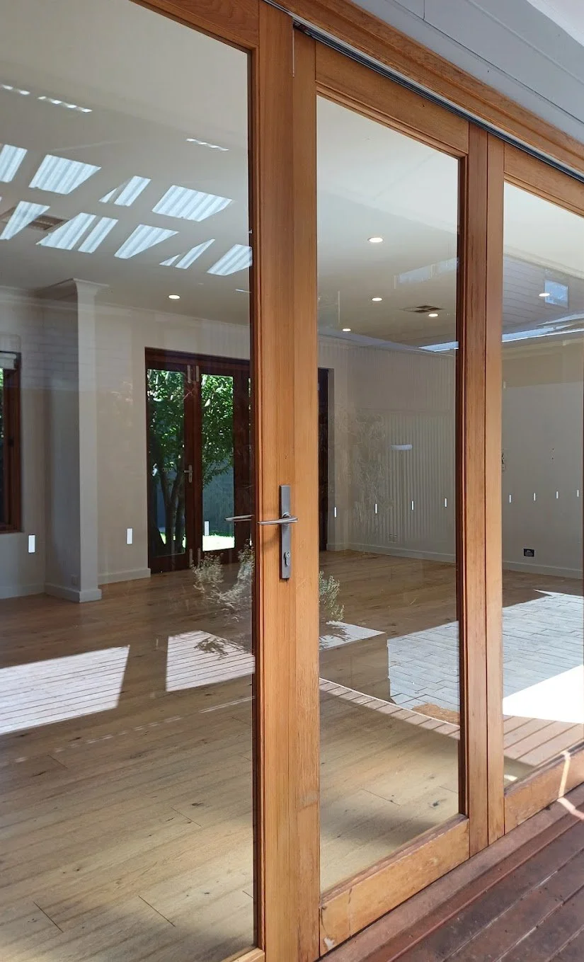 Interior view of a room with glass sliding doors, wooden frames, hardwood flooring, and recessed ceiling lights, with outdoor greenery visible through the glass.