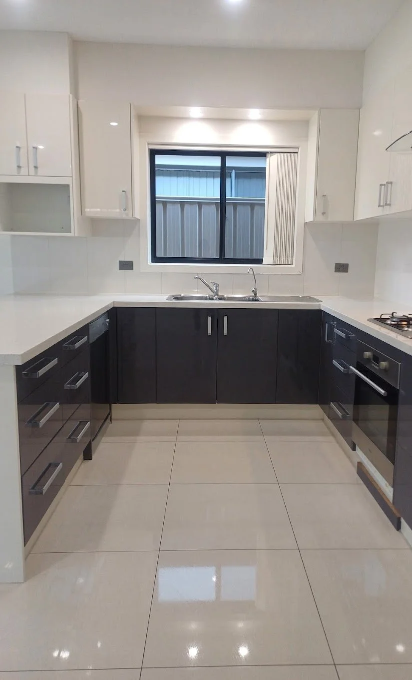 Modern kitchen with white upper cabinets, black lower cabinets, white countertops, a window above the sink, tiled floor, and built-in oven and stovetop.
