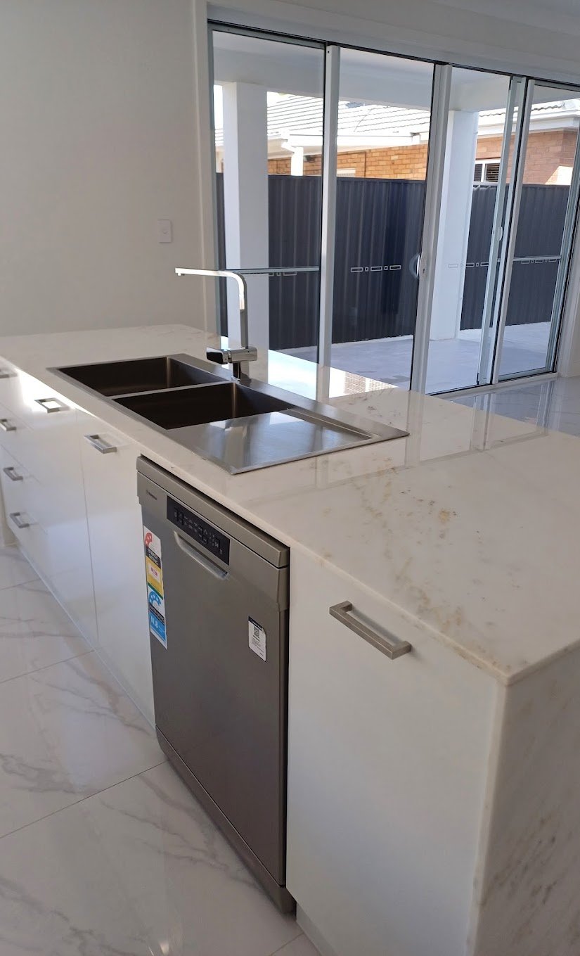 Modern kitchen with a double sink and a dishwasher beneath marble countertop, overlooking a sliding glass door leading to an outdoor area.