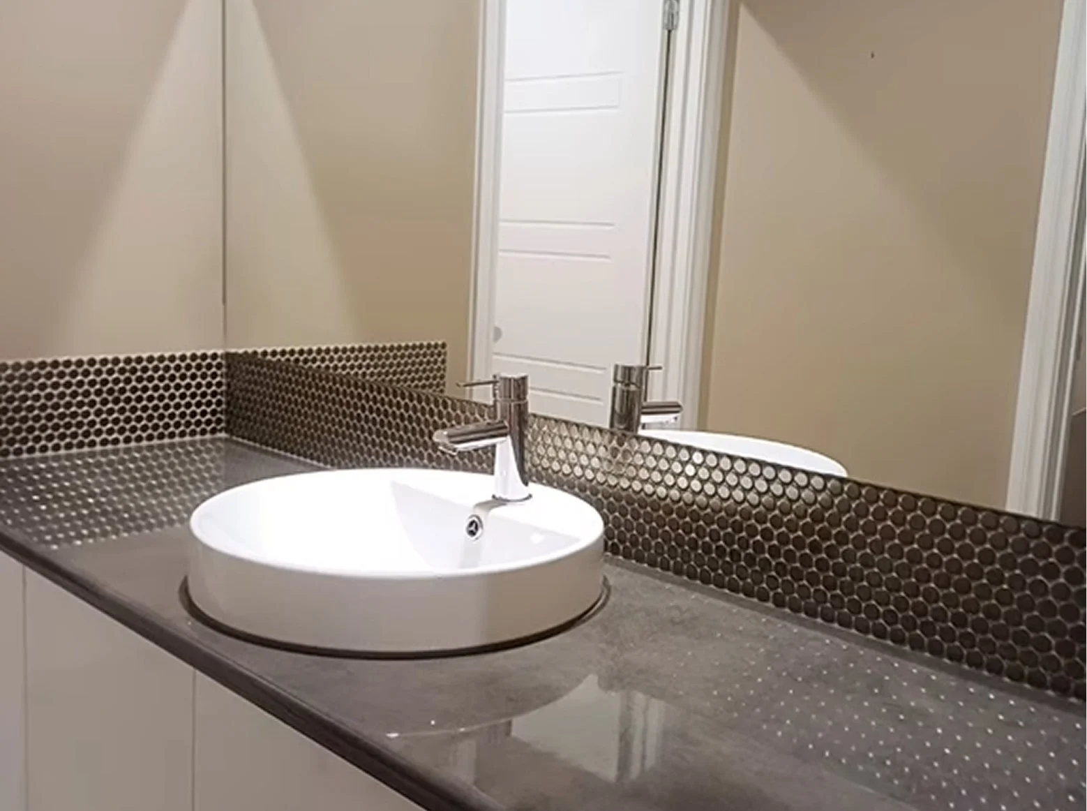 Modern bathroom sink with a round vessel basin on a dark countertop, with a mosaic tile backsplash, a mirror, and a door reflected in the mirror.