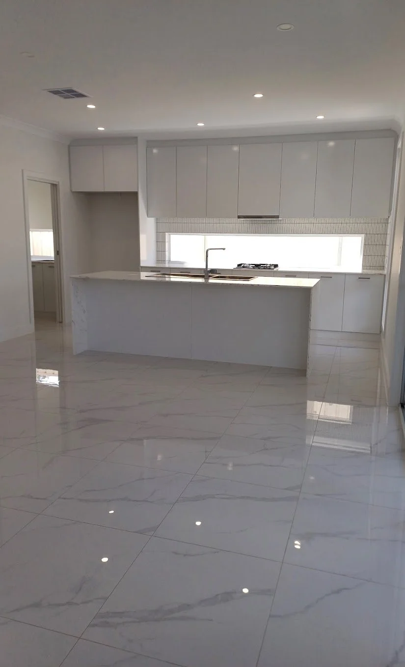 Empty modern kitchen with white cabinets, marble island, and glossy white tile flooring.