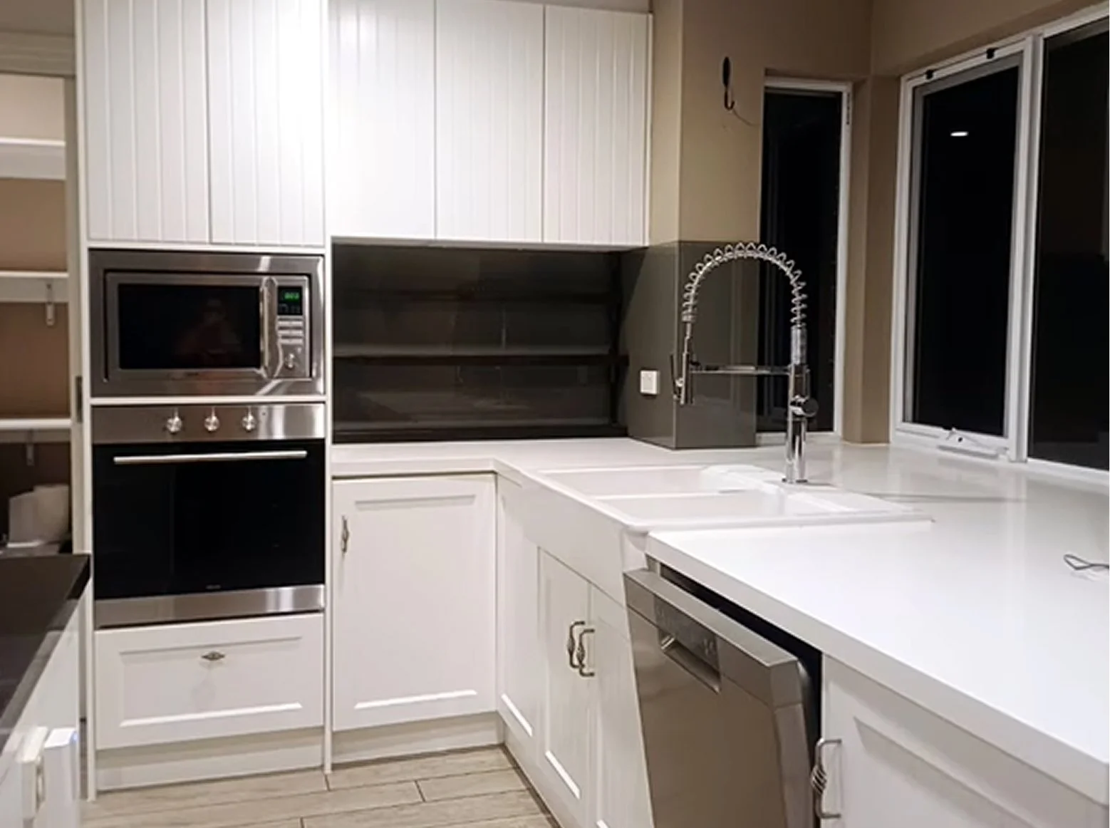 Modern kitchen with white cabinets, stainless steel appliances, a double basin sink, and a black open shelving unit, illuminated by a window and warm lighting.