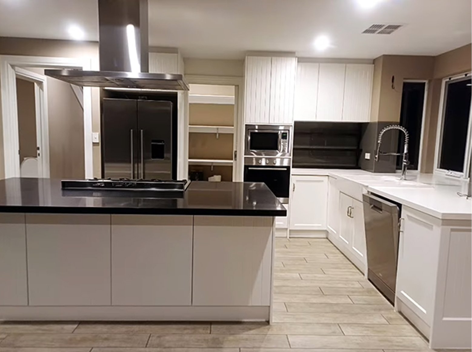 Kitchen with white cabinets, black countertops, stainless steel appliances, and a black open shelving unit. There is a large window over the sink and a kitchen island in the foreground.