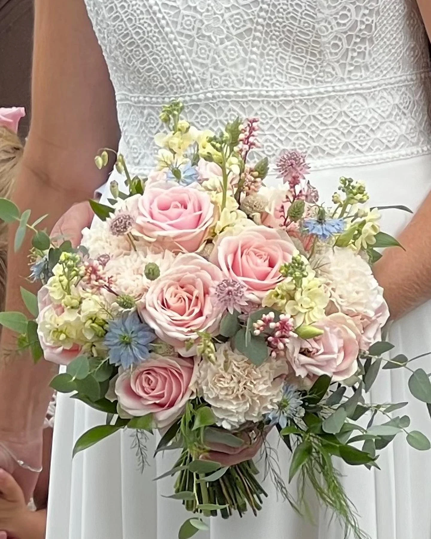 En bukett med ljusrosa rosor, vita och blå blommor, gröna blad, och andra småblommor, hålls av en person i vit klänning.