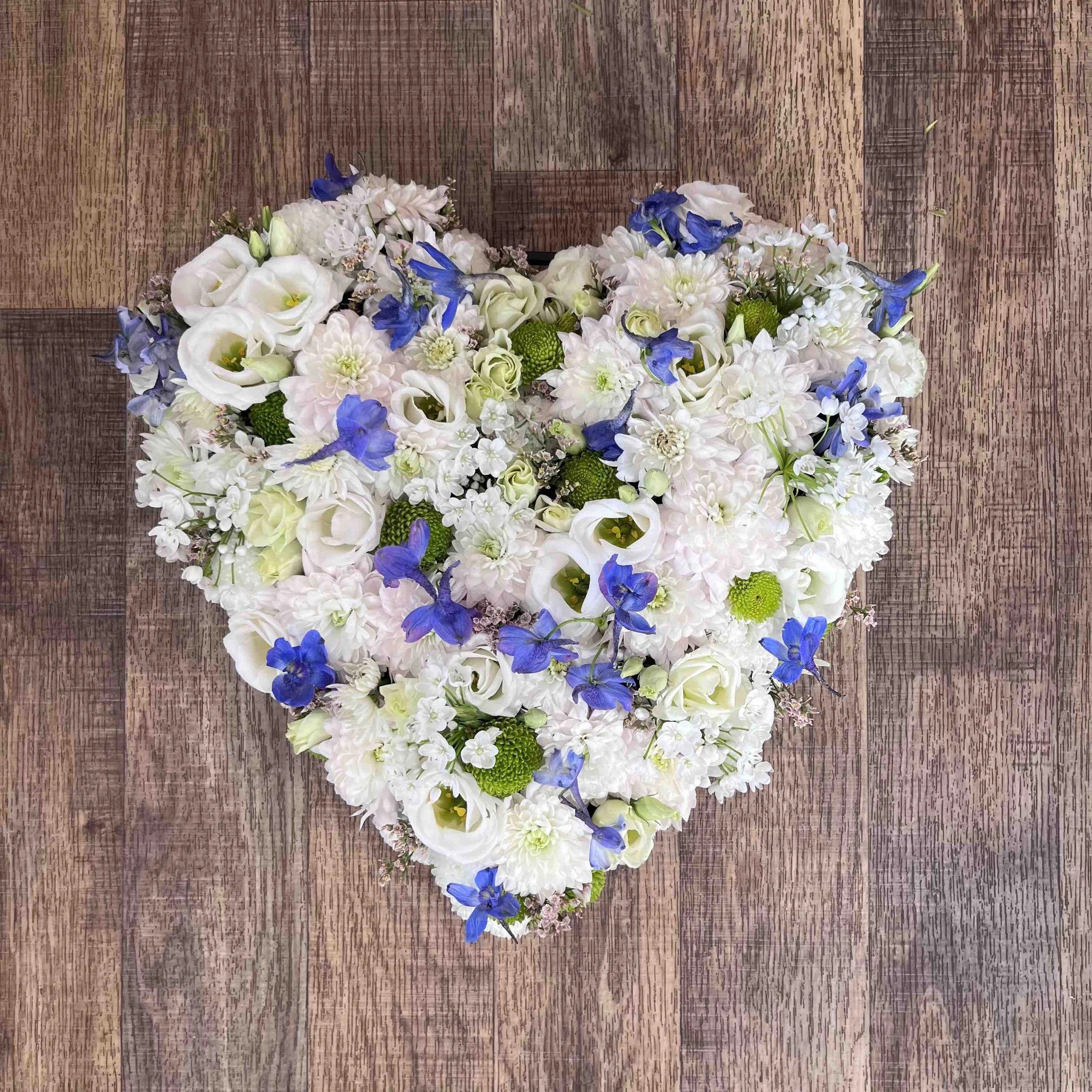 En blomsterarrangemang i form av ett hjärta med vita, blå och gröna blommor på ett trägolv.
