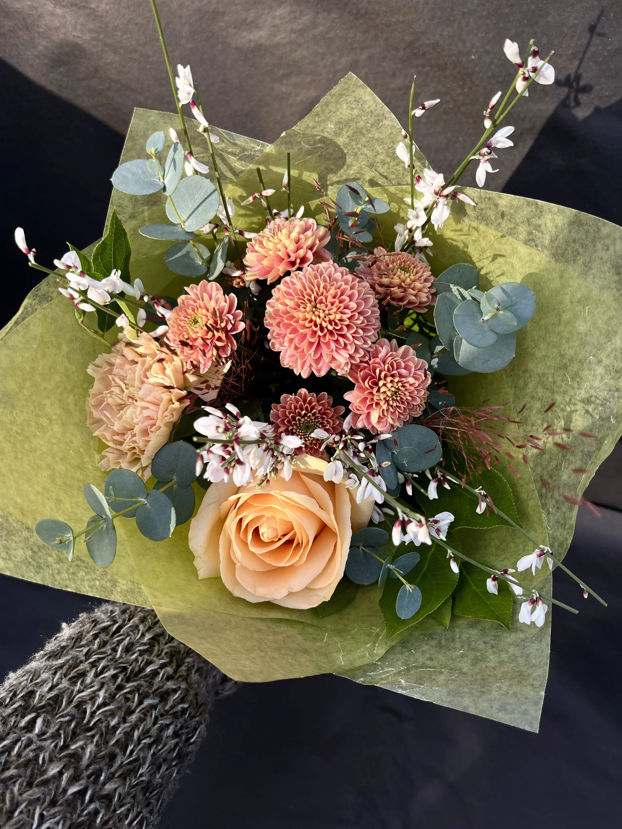 Blomsterbukett med rosa och persikofärgade blommor, gröna blad och små vita blommor, omsluten av ljusgrönt silkespapper