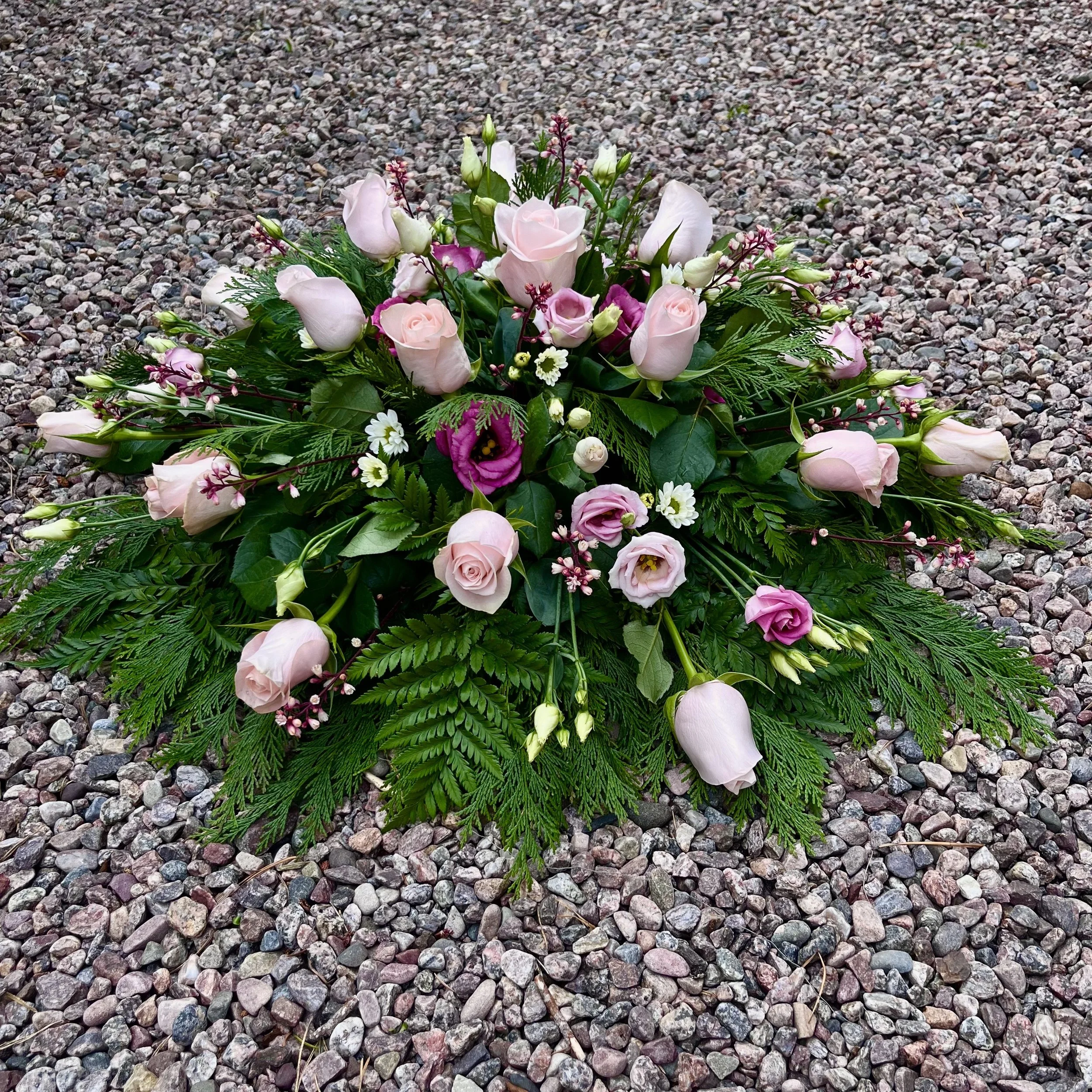Väldigt vacker begravningsdekoration med ljusrosa rosor och mörkrosa blommor samt gröna blad, placerad på grusbeläggning i utomhusmiljö.