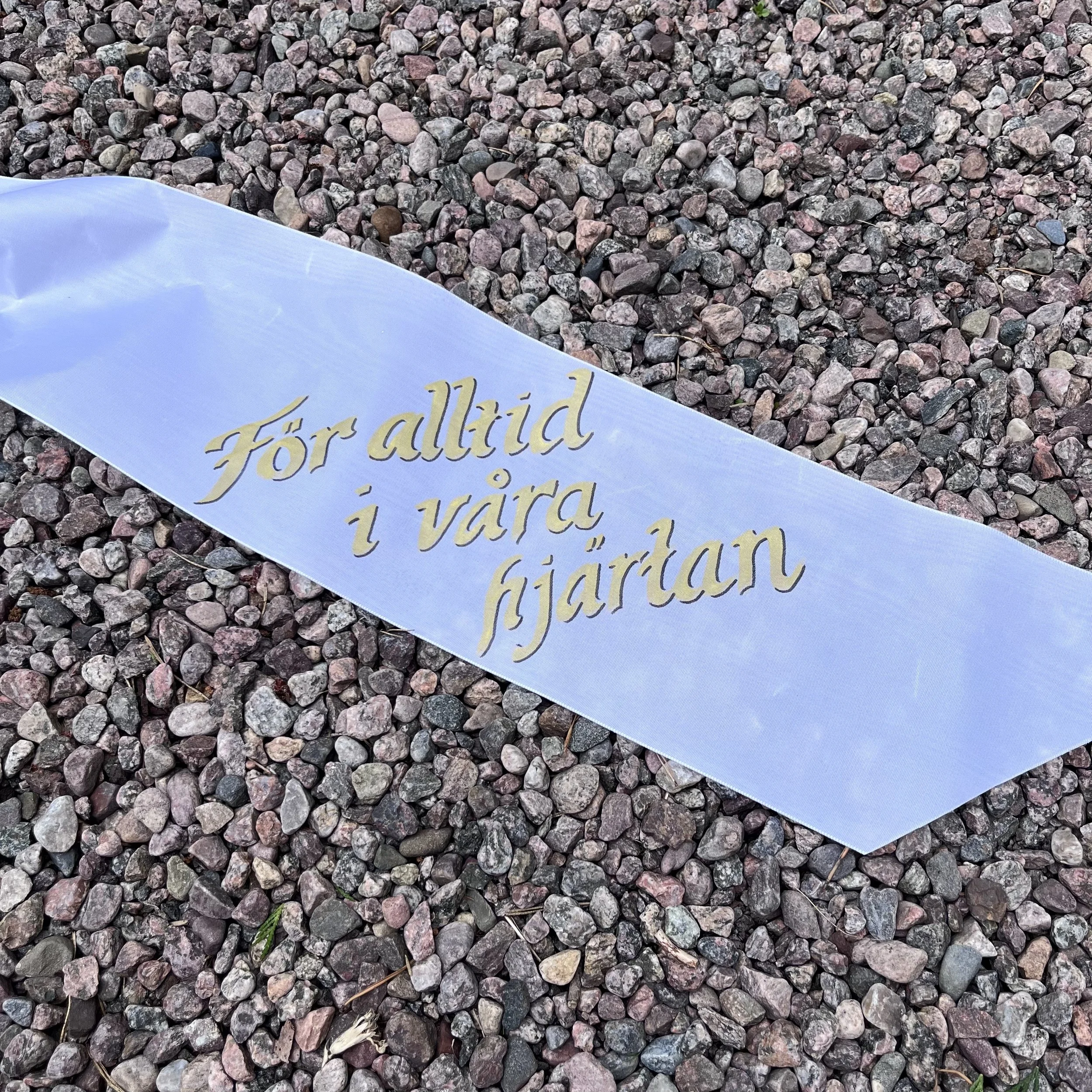 En vit kransband med guldfärgad text på som säger 'För alltid i våra hjärtan' ligger på en yta av smågrus.