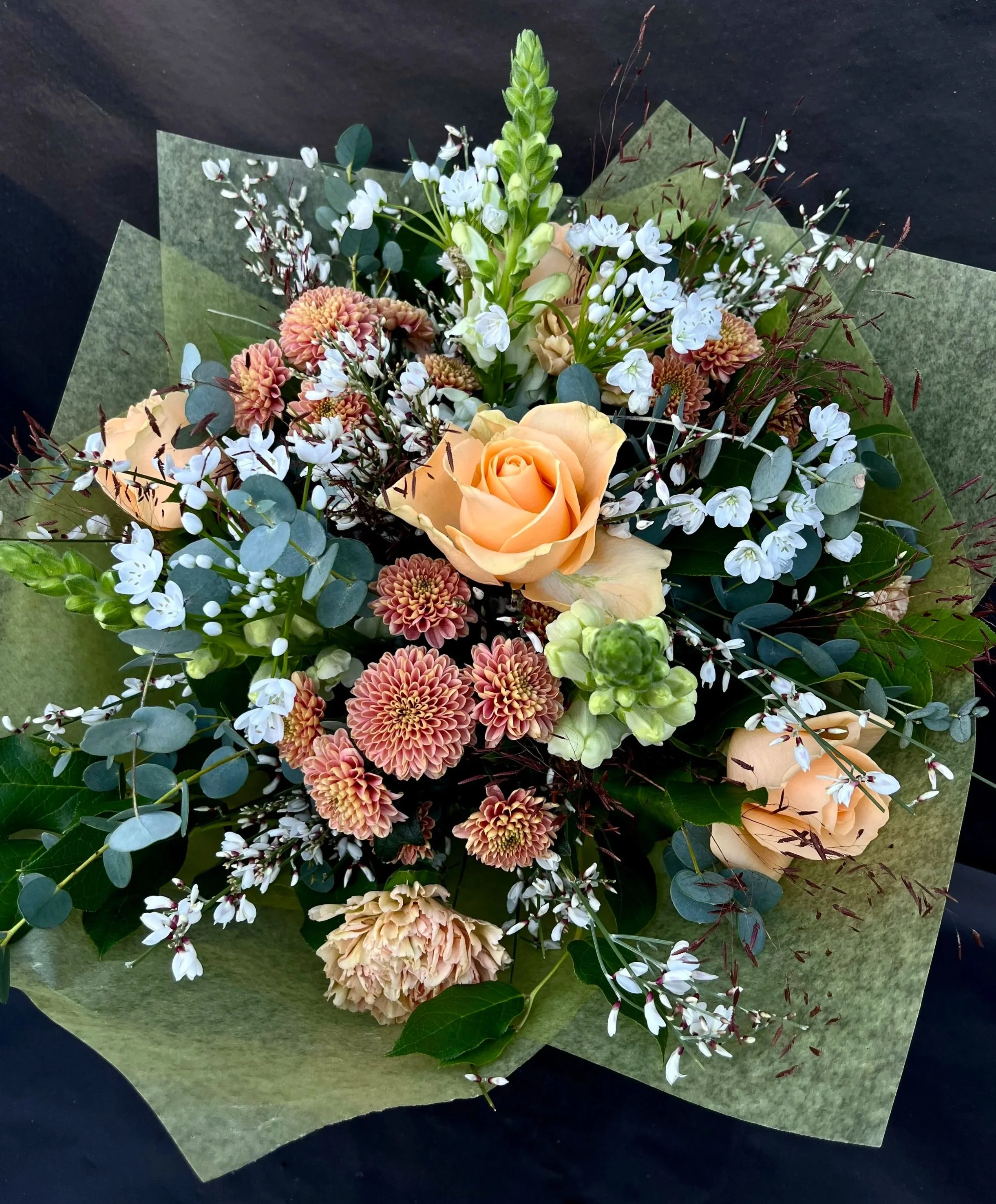 Blommor i en stor bukett med aprikos ros, vit ginst, chrystanthemum och gröna blad omsluten av grönt silkespapper.