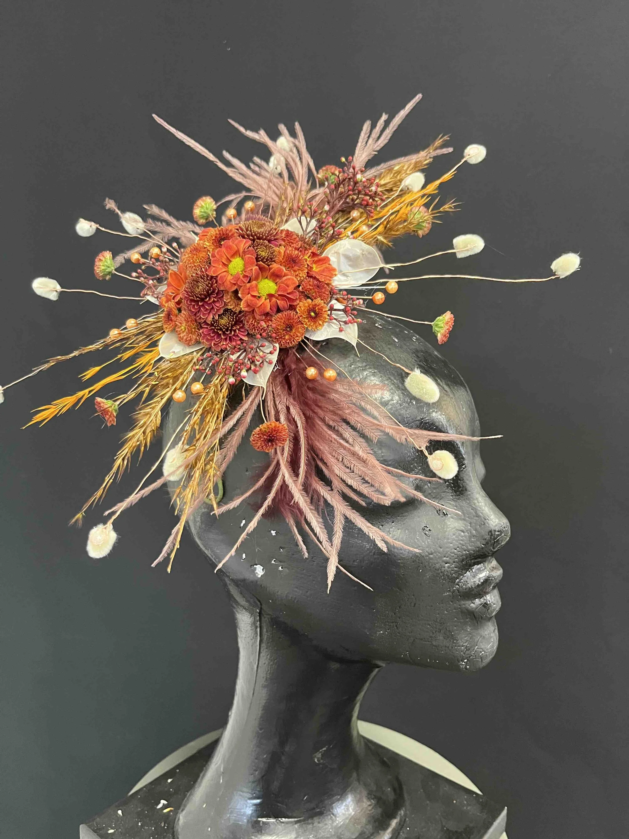 En svart konstgjord huvudstaty med ett färgstarkt blomsterarrangemang på huvudet, inklusive röda, orangea och gula blommor och torkade växter, mot en svart bakgrund.