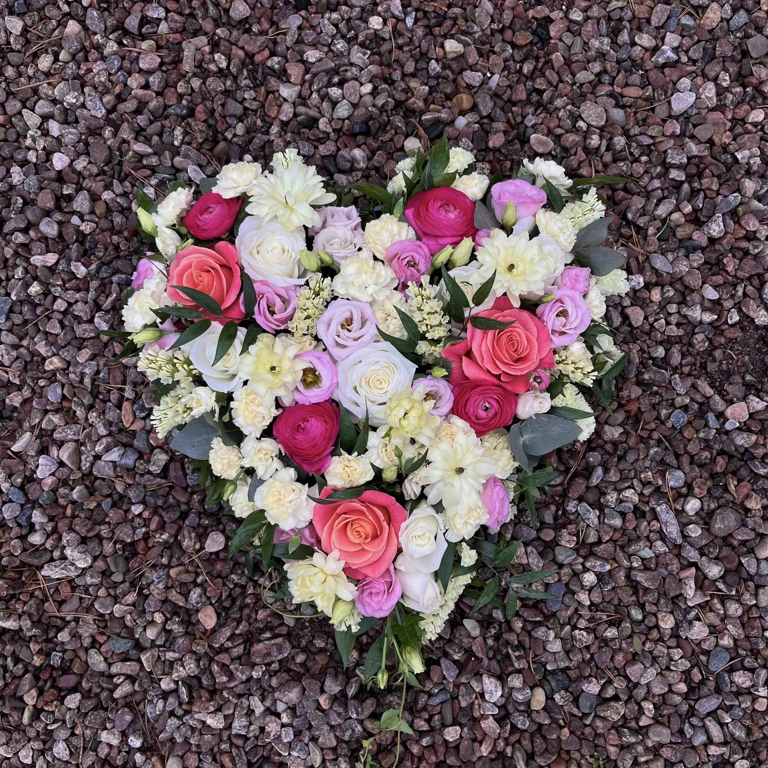 En blomsterdekoration formad som ett hjärta, placerad på en grusig yta. Buketten innehåller rosa, vita, lavendel och gula blommor.
