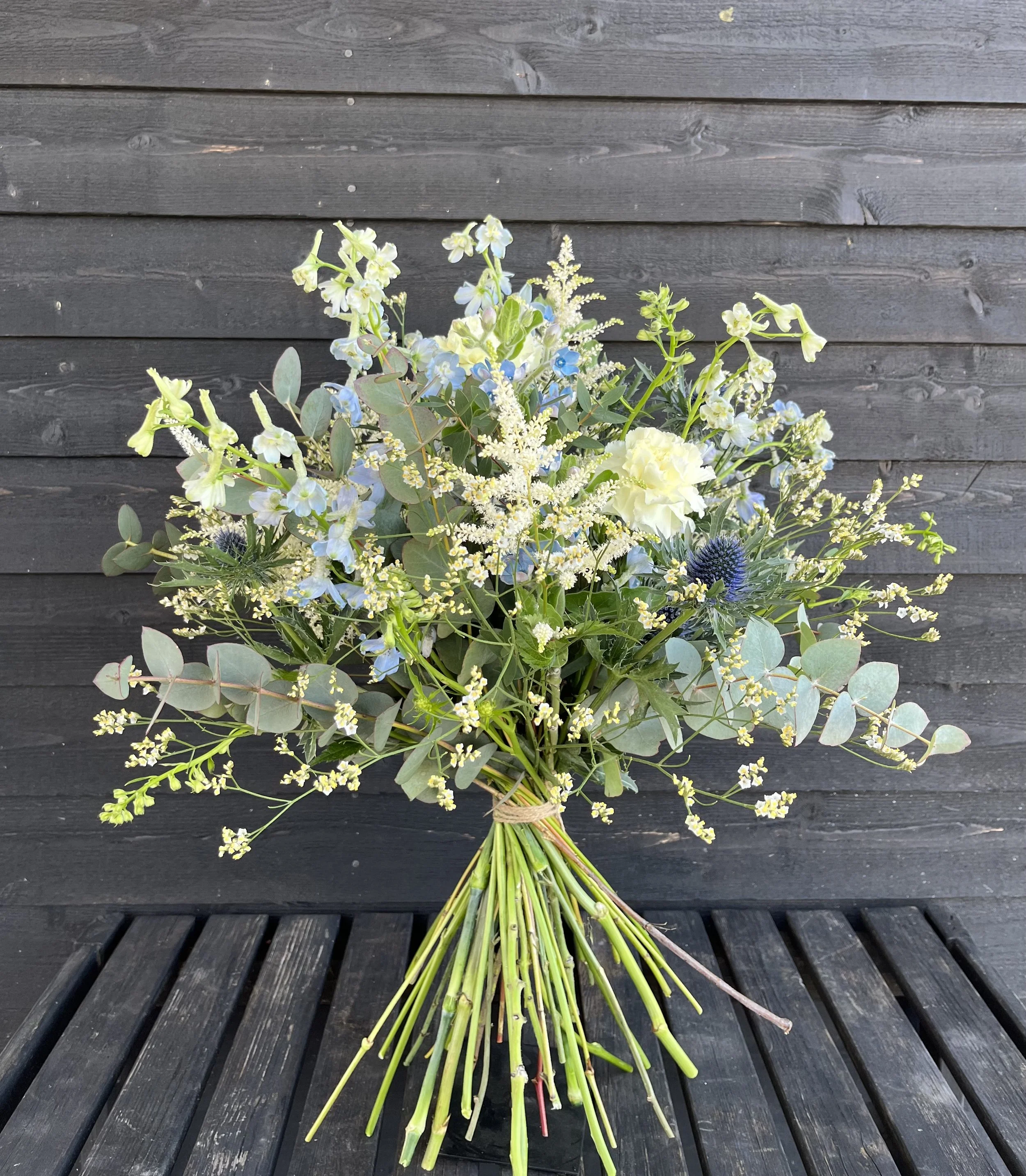 En bukett med vita, blå och gröna blommor, sammanbunden med ett snöre, placerad på ett svart bord mot en mörk trävägg.