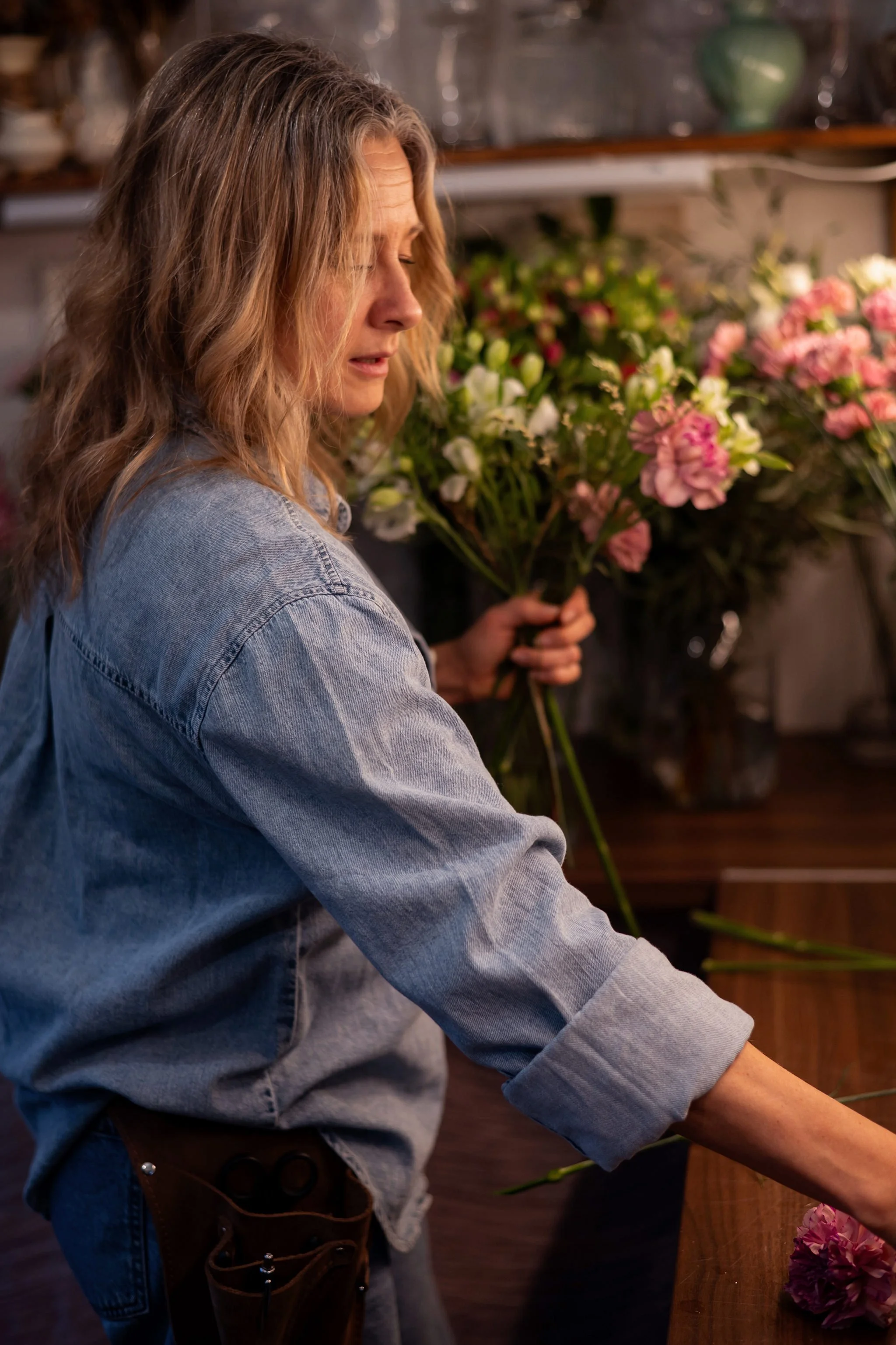 Kvinna i denimjacka som arrangerar snittblommor i ett blomsterbutik.