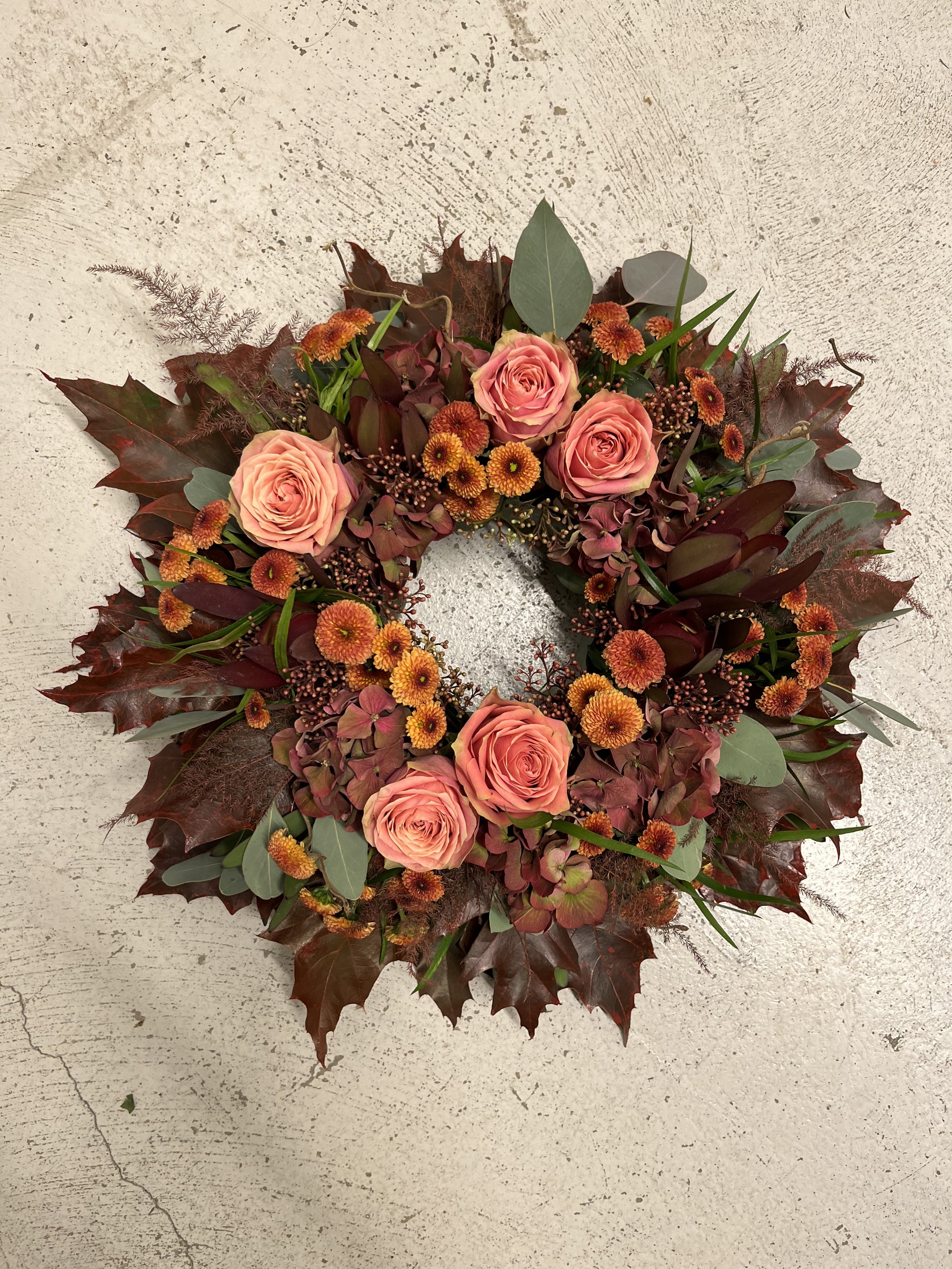 En blomsterarrangemang i form av en krans med aprokosa rosor, orange och gula blommor, eukalyptusblad och mörkröda löv.