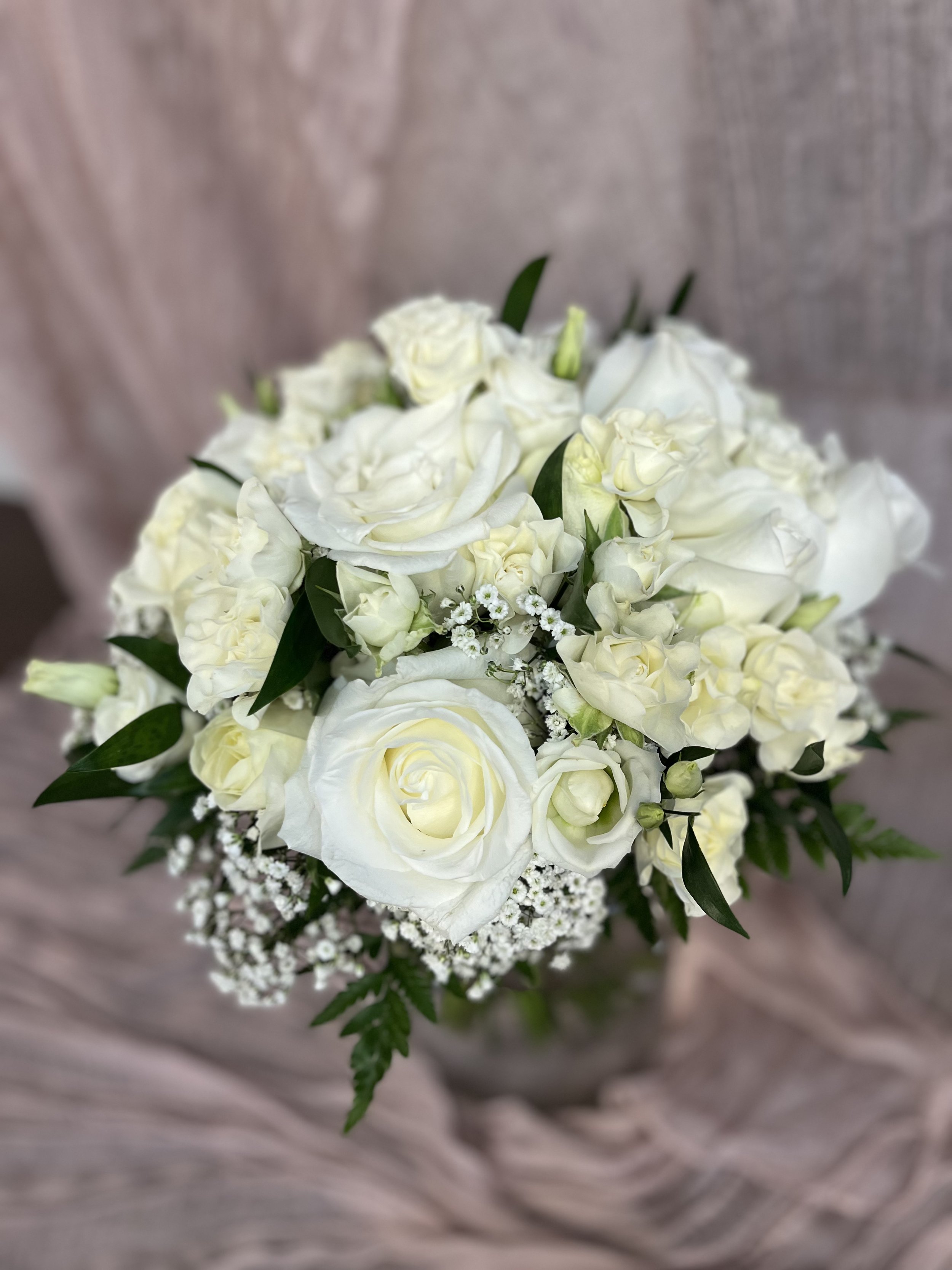 En bukett av vita blommor, inklusive rosor, liljor och mindre blomster, med gröna blad, mot en ljus bakgrund.